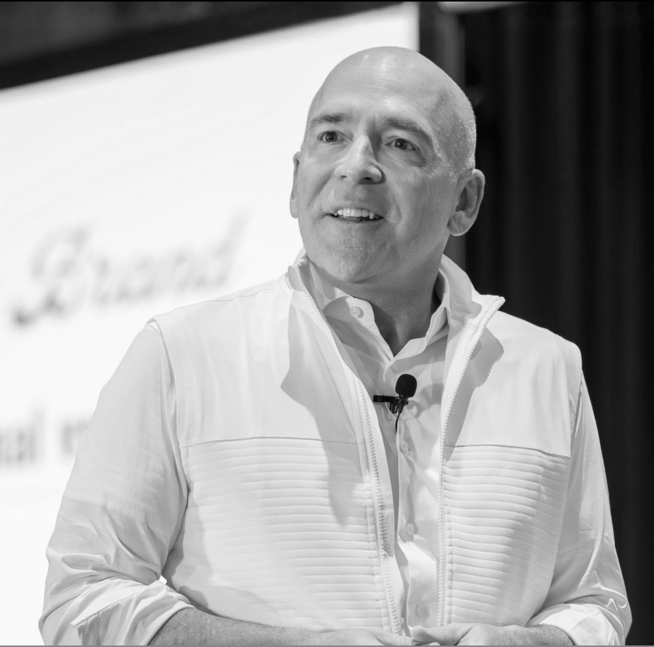 A black and white photo of a man with a bald head, wearing a white jacket and dress shirt, speaking at an event with a microphone attached to his shirt.