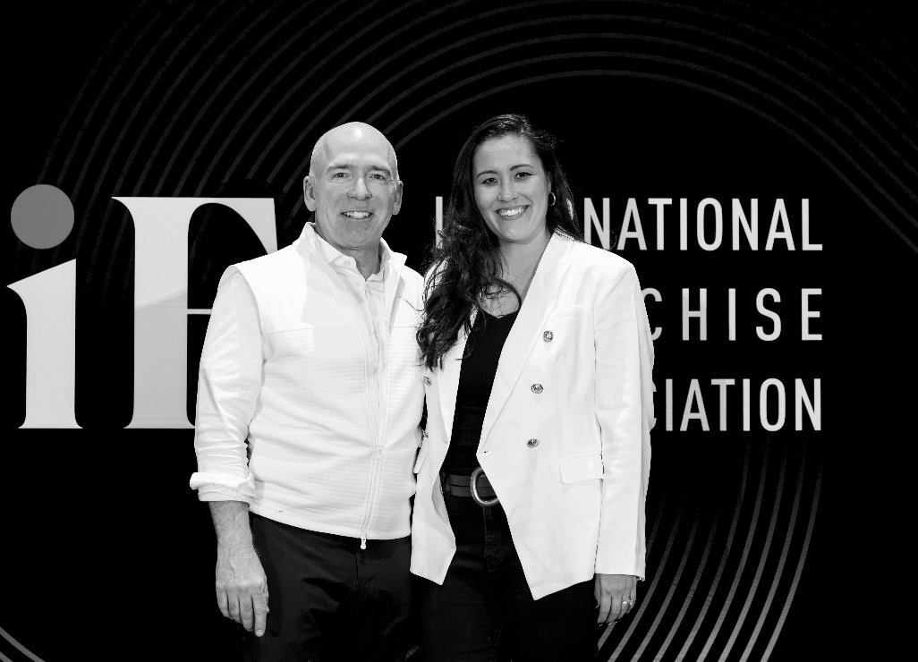 Two people smiling at an event with a large sign in the background reading 'American International Franchise Association'.