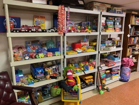 A white bookshelf filled with various toys and games, including trucks, dolls, and board games, in a room with a brown carpet.