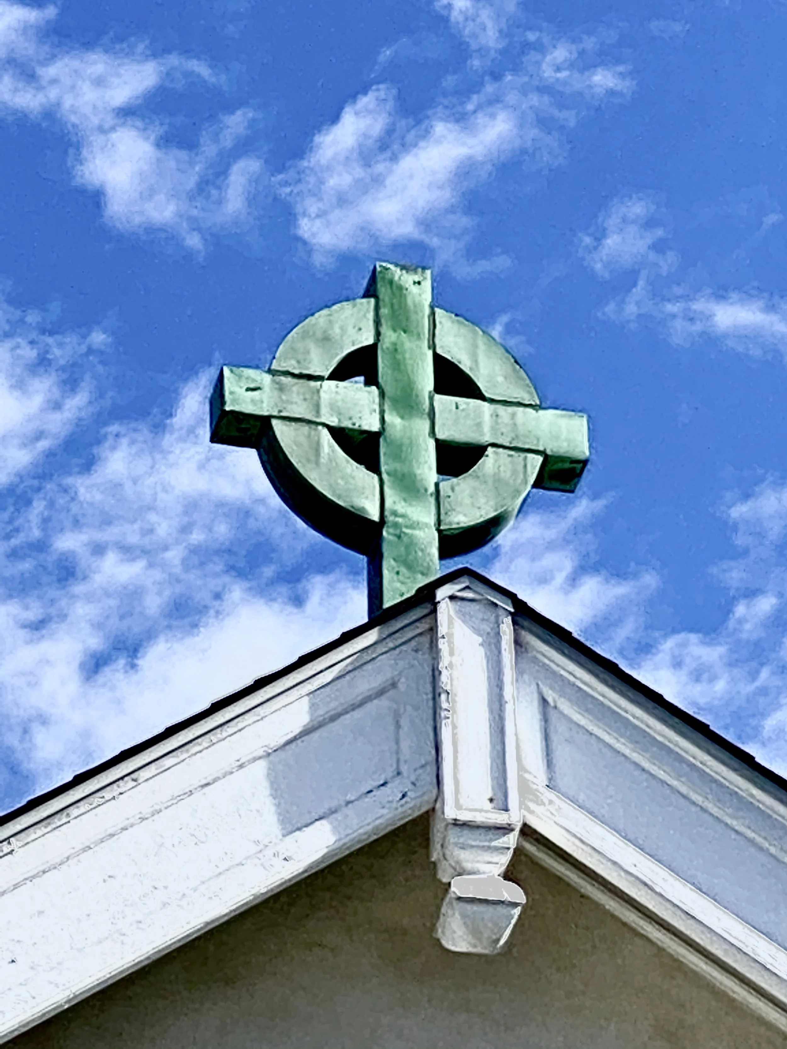 Green Celtic cross weather vane on the roof corner against blue sky with clouds.