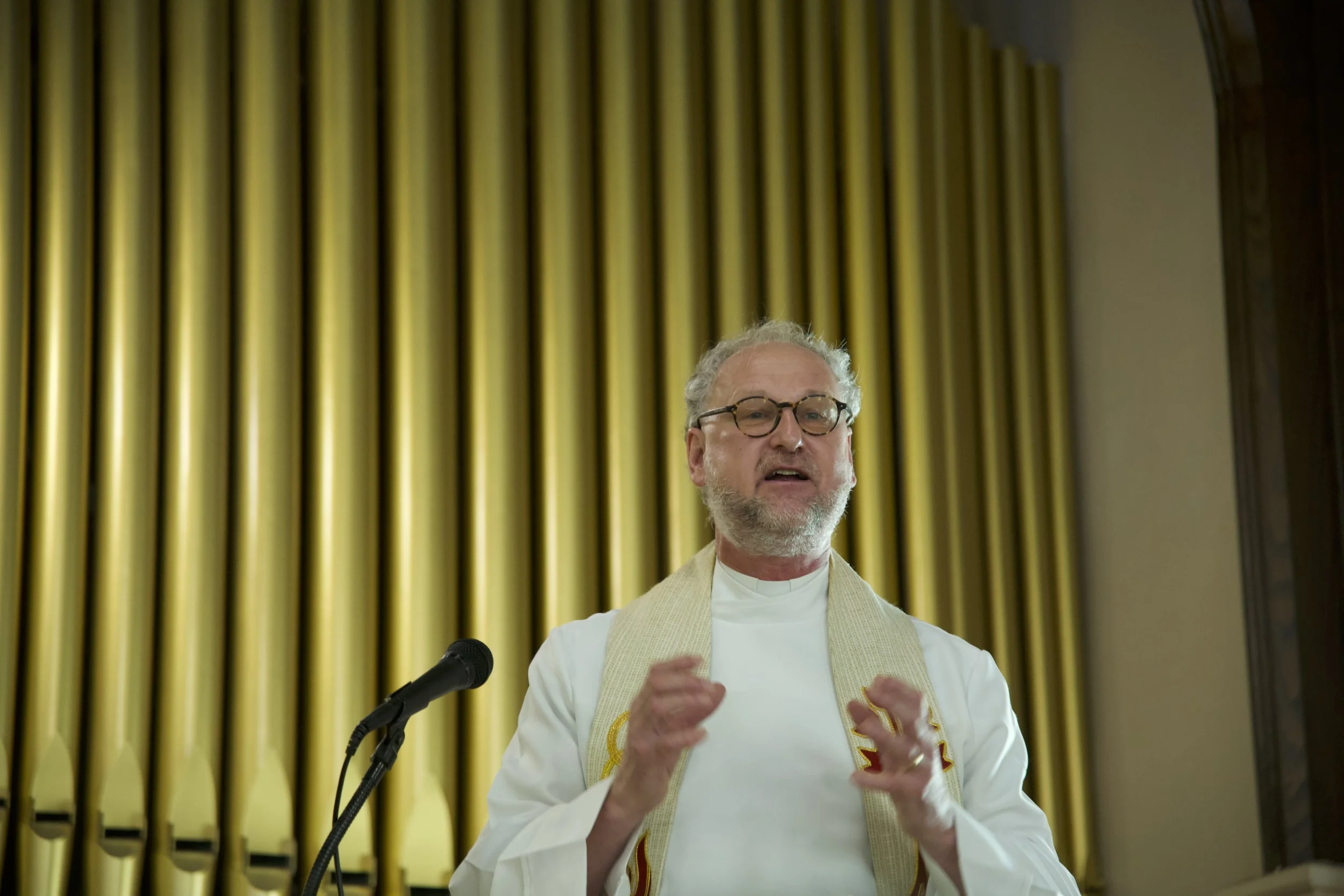 A man with glasses and gray hair speaking at a microphone in front of large gold-colored pipes.