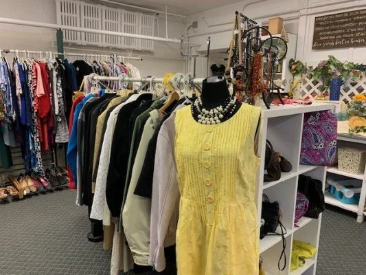 Clothing rack with various jackets, shirts, and dresses, a yellow dress on a mannequin, and jewelry on display inside a thrift store or boutique.