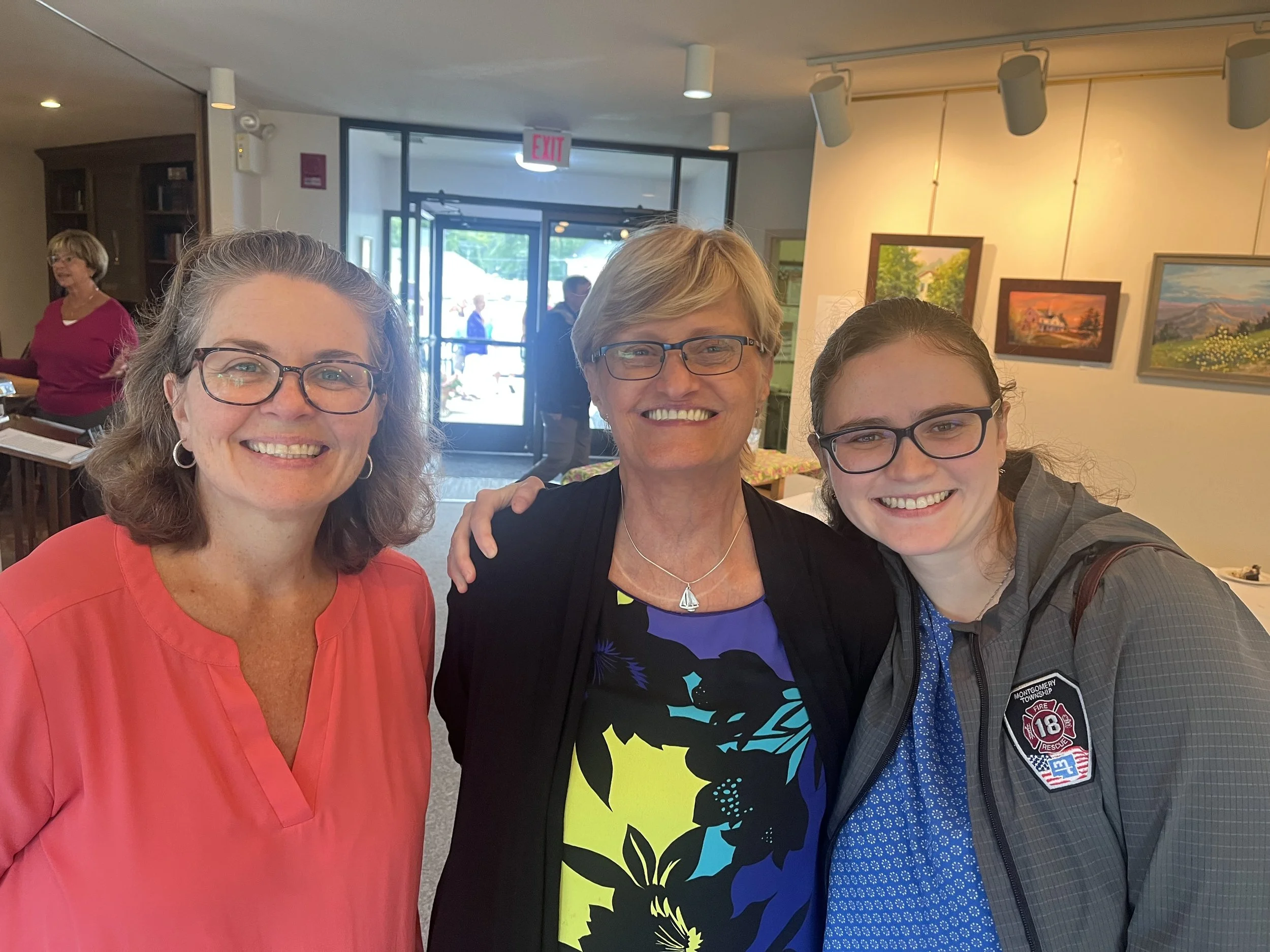 Three women standing together smiling indoors at an art gallery.