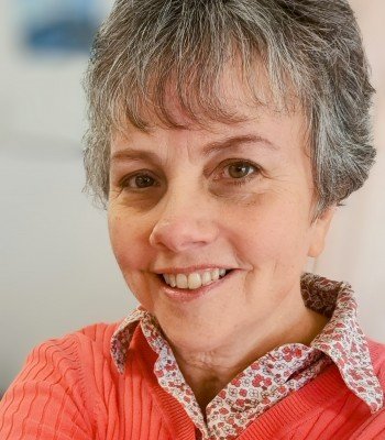 A woman with short gray hair smiling at the camera, wearing a red sweater and a patterned collared shirt.