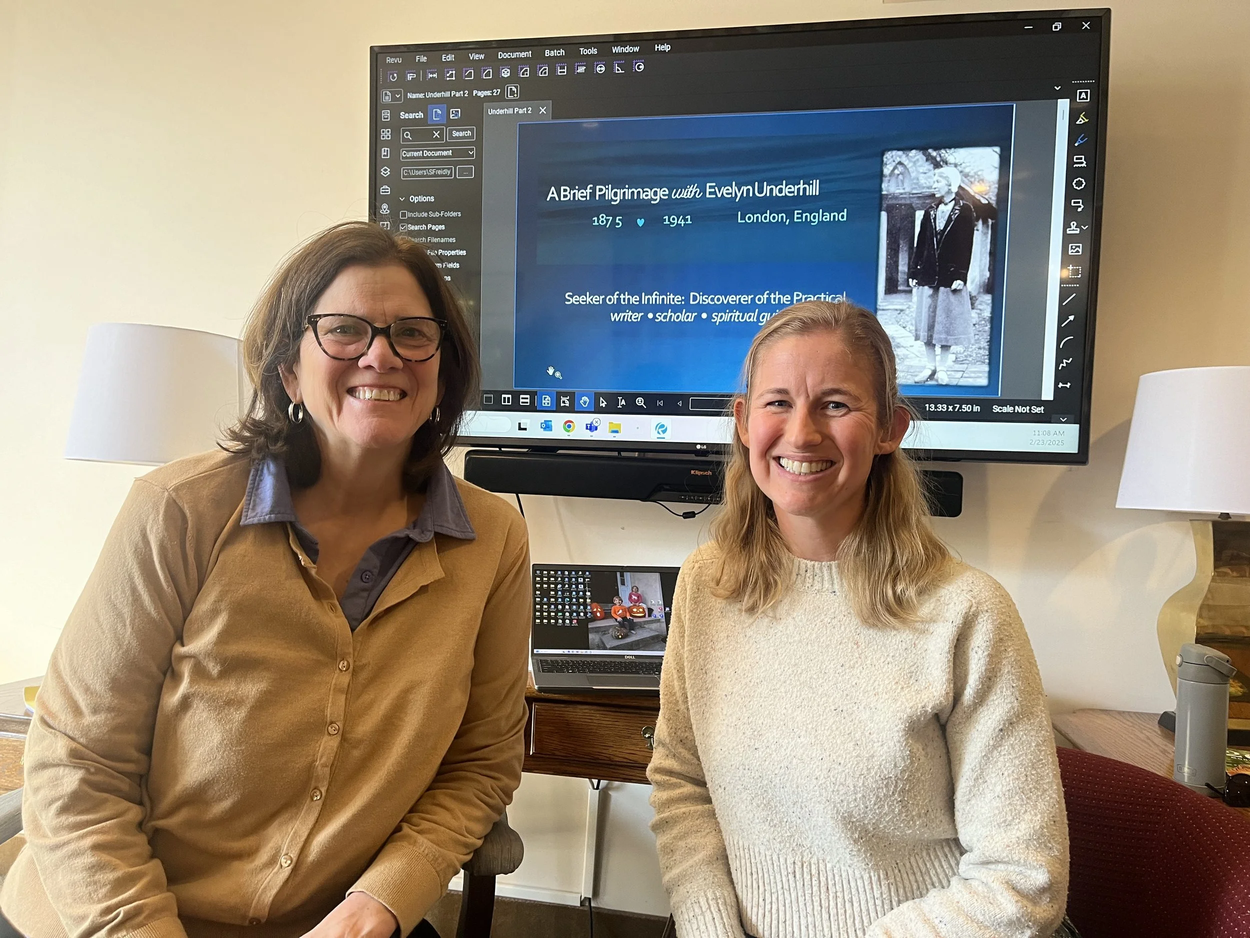 Two women smiling and sitting in front of a large monitor displaying a presentation slide about Evelyn Underhill.