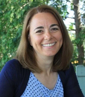 A woman with shoulder-length brown hair, smiling outdoors with trees and sunlight in the background.