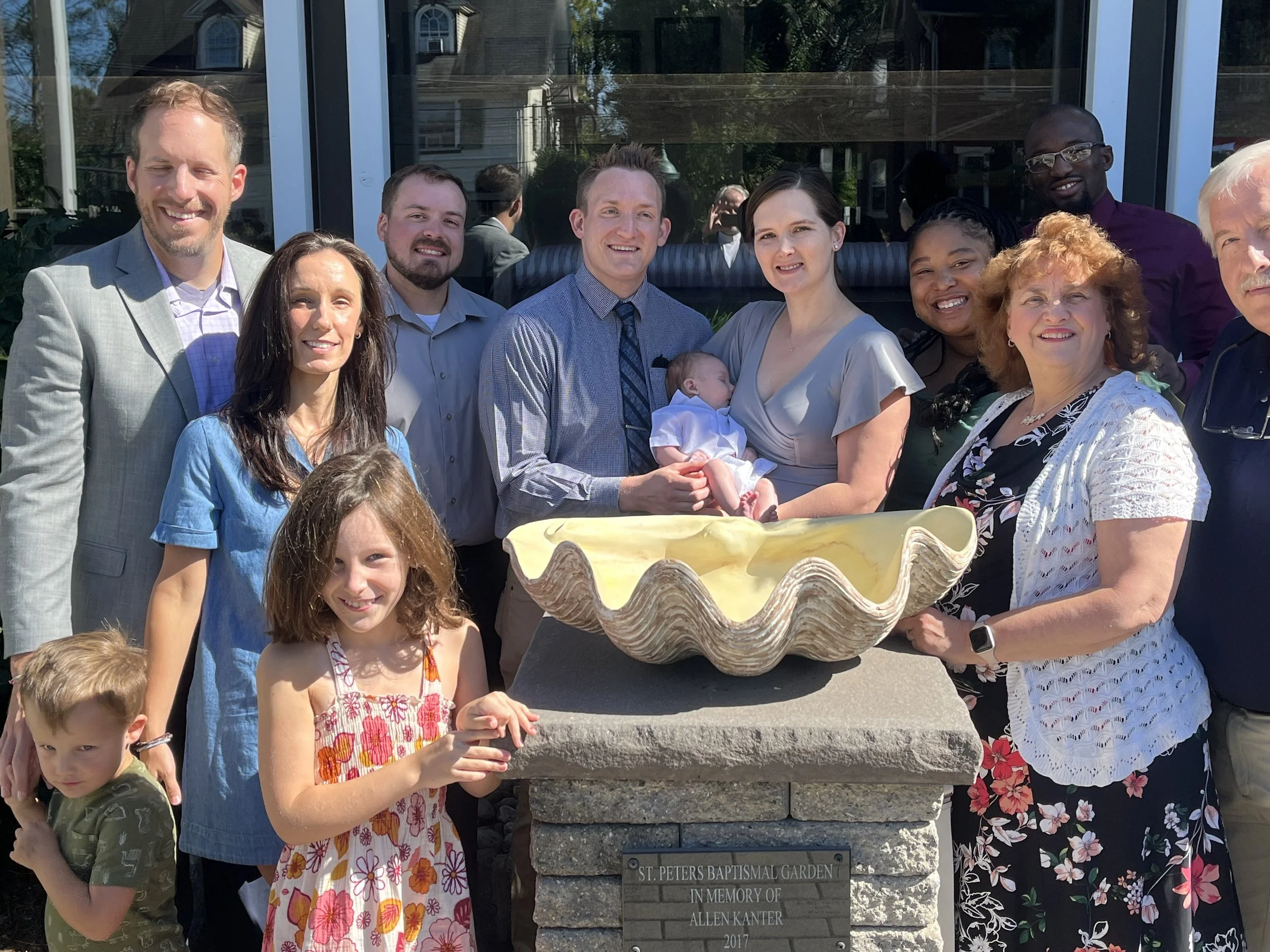 Family and friends gathered around a memorial fountain at the St. Peters Baptismal Garden, celebrating a baptism, with everyone smiling in bright sunlight.