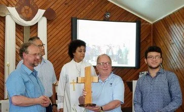 A group of five people, including a man holding a wooden cross and a woman holding a Bible, standing in a room with wooden paneling and a projector screen behind them. The woman is wearing a white robe with a gold cross.