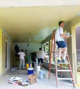 Group of people painting walls inside a house, with one person on a ladder applying paint