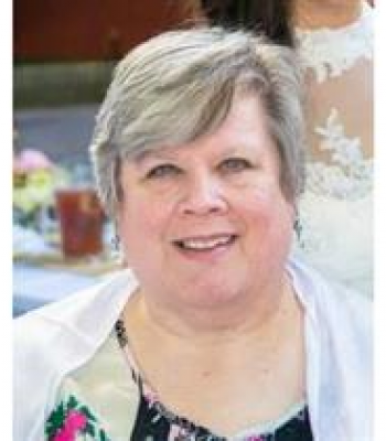 A smiling woman with short gray hair, wearing a white jacket and a floral top, at an outdoor gathering.