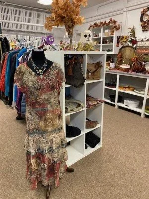A vintage clothing store displays a mannequin dressed in a patchwork dress, with shelving units behind holding handbags, decor, and accessories; floral arrangements and decorative skulls are on top of the shelves.