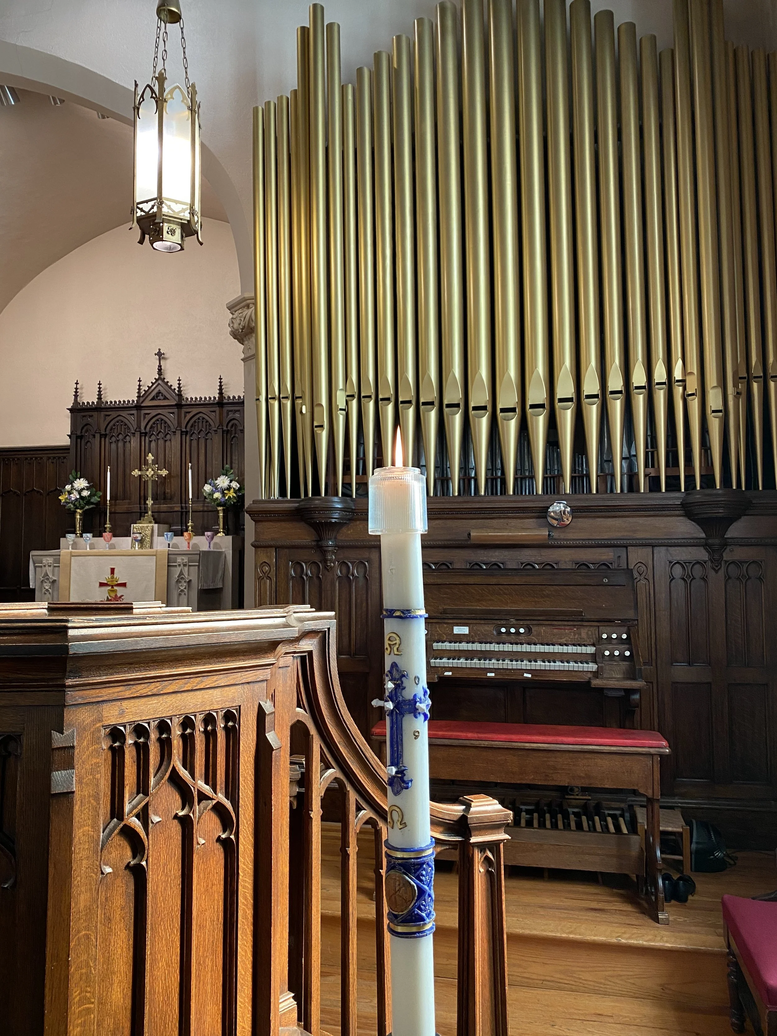 A lit candle with blue and gold decorations is placed on a wooden railing inside a church. Behind it, there is an organ with golden pipes, an altar with flowers, candlesticks, and a cross. The church has wooden pews and a high, arched ceiling with hanging chandeliers.