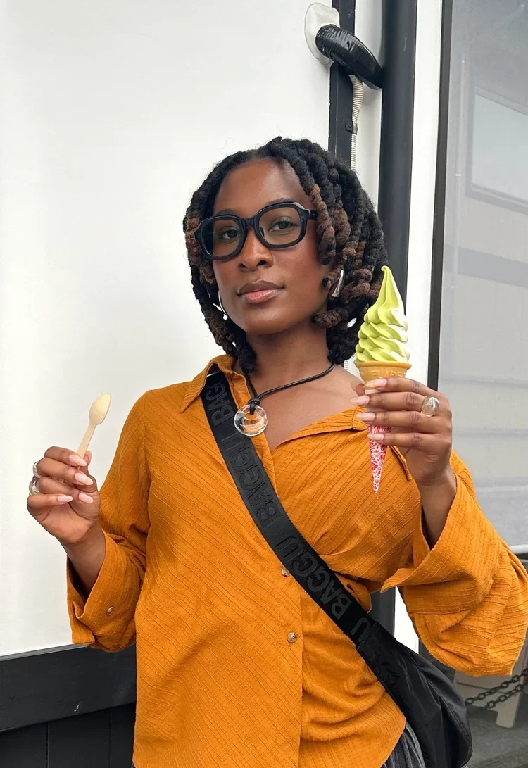 A woman with glasses and braided hair holding a yellow ice cream cone in one hand and a small wooden spoon in the other, standing indoors against a plain background.