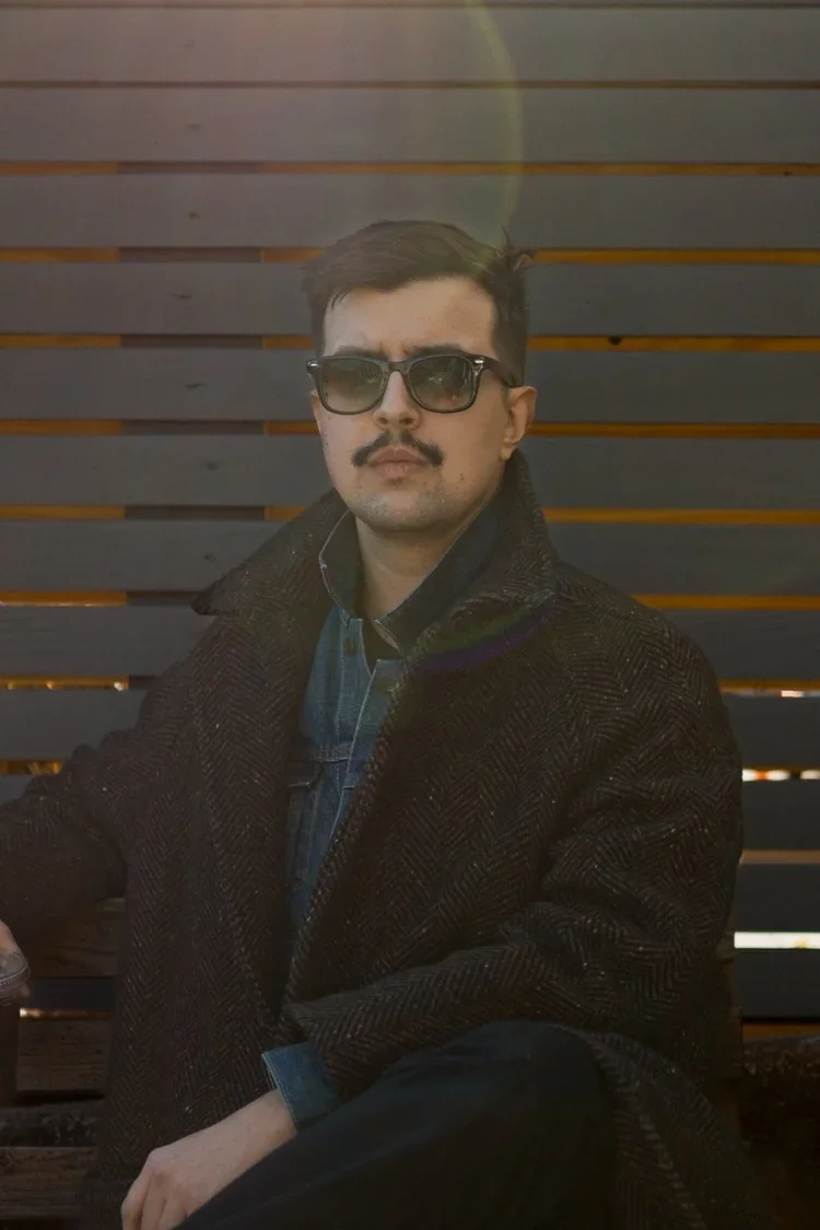 A man with dark hair, a mustache, wearing sunglasses, a dark coat, and a denim shirt sitting in front of a wooden wall.