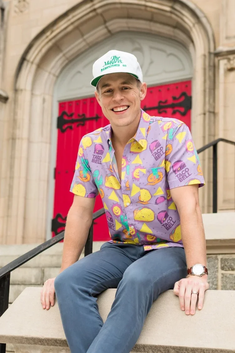 A young man sitting on stairs in front of a red door with black decorative hinges. He is smiling, wearing a colorful Taco Bell themed shirt, a white cap with green text, a wristwatch, and a wedding ring.