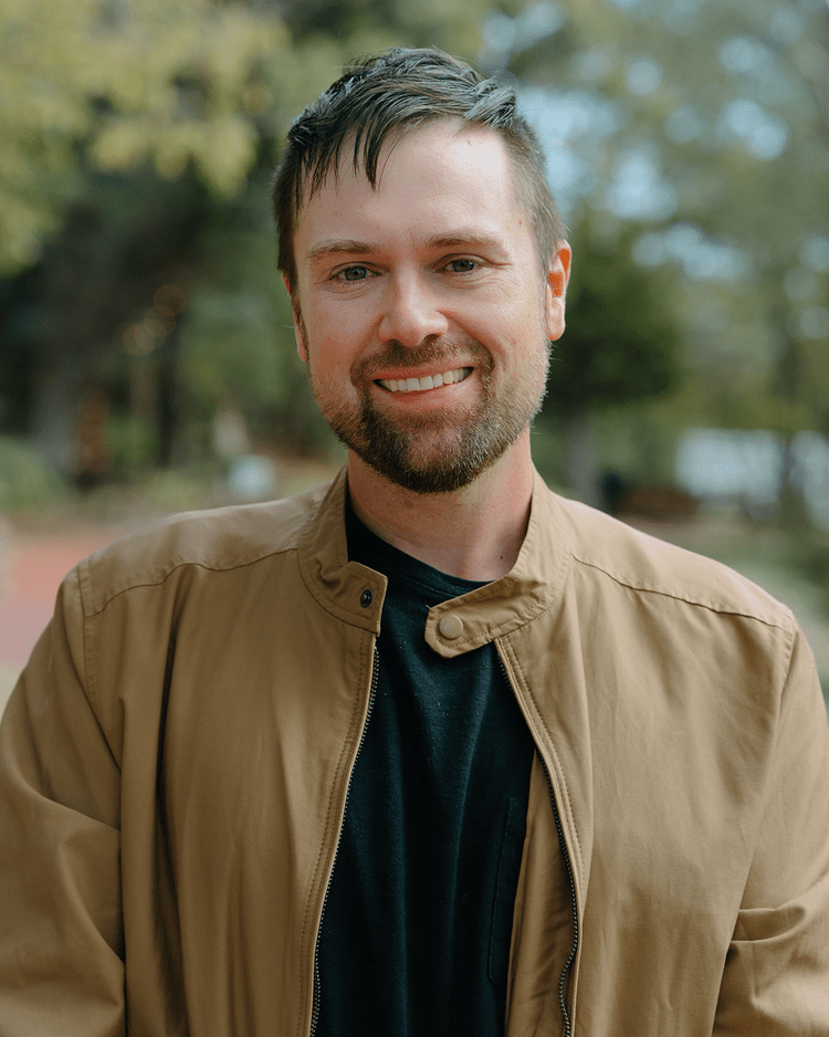 A smiling man with short, dark hair and a beard, wearing a tan jacket and black shirt, outdoors with blurred trees in the background.