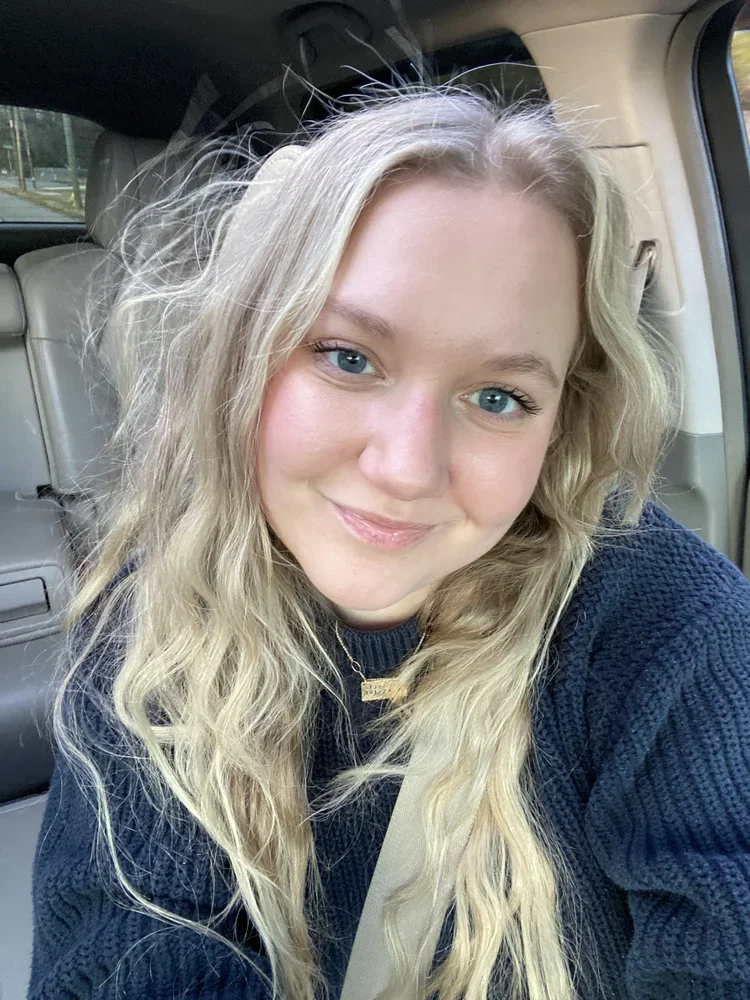 A young woman with long wavy blonde hair and blue eyes smiling while sitting in the passenger seat of a car, wearing a black sweater and a gold necklace.