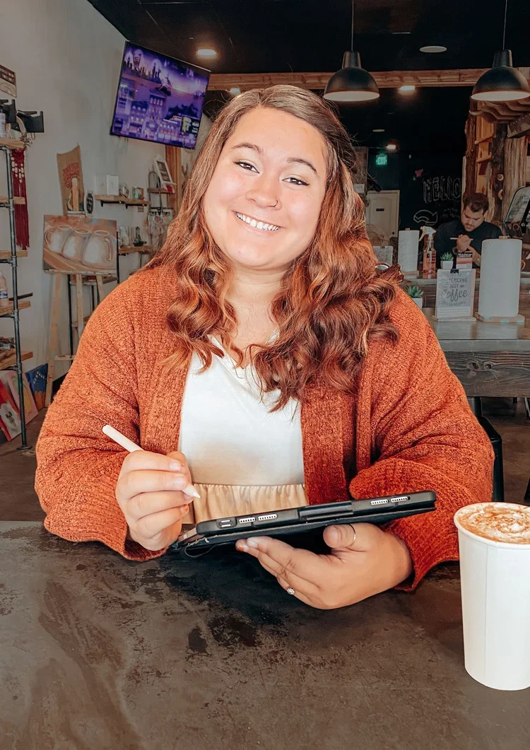 A smiling woman with curly red hair sitting at a table in a coffee shop, holding a tablet and stylus, with a coffee cup in front of her. The background shows a TV, shelves with books and decorations, and a person working behind the counter.