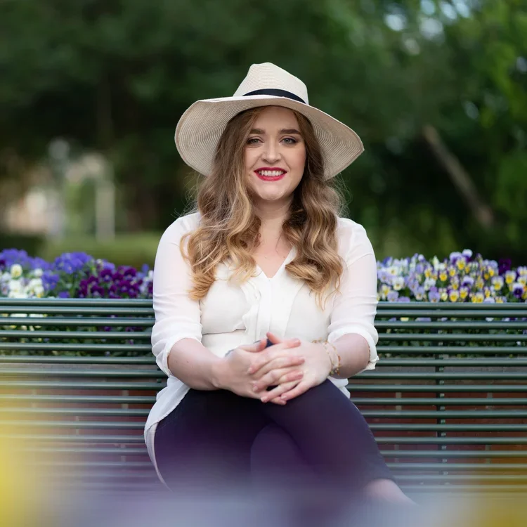 Woman with long, wavy hair wearing a white hat, white blouse, and black pants, sitting on a park bench with purple and yellow flowers in the background.