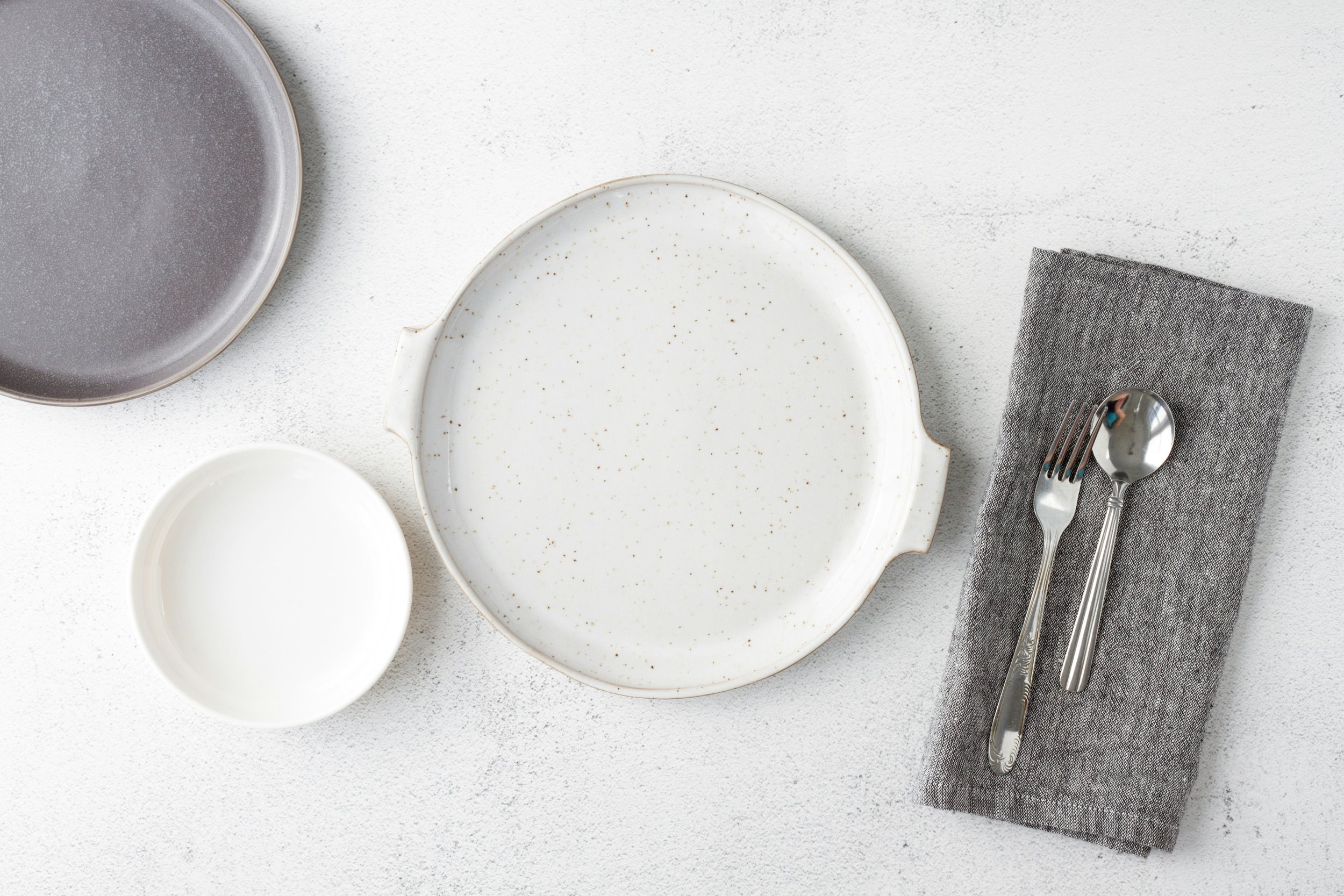Empty white ceramic plate, small white bowl, gray ceramic dish, gray napkin with silver fork and spoon on top, all on a white textured surface.