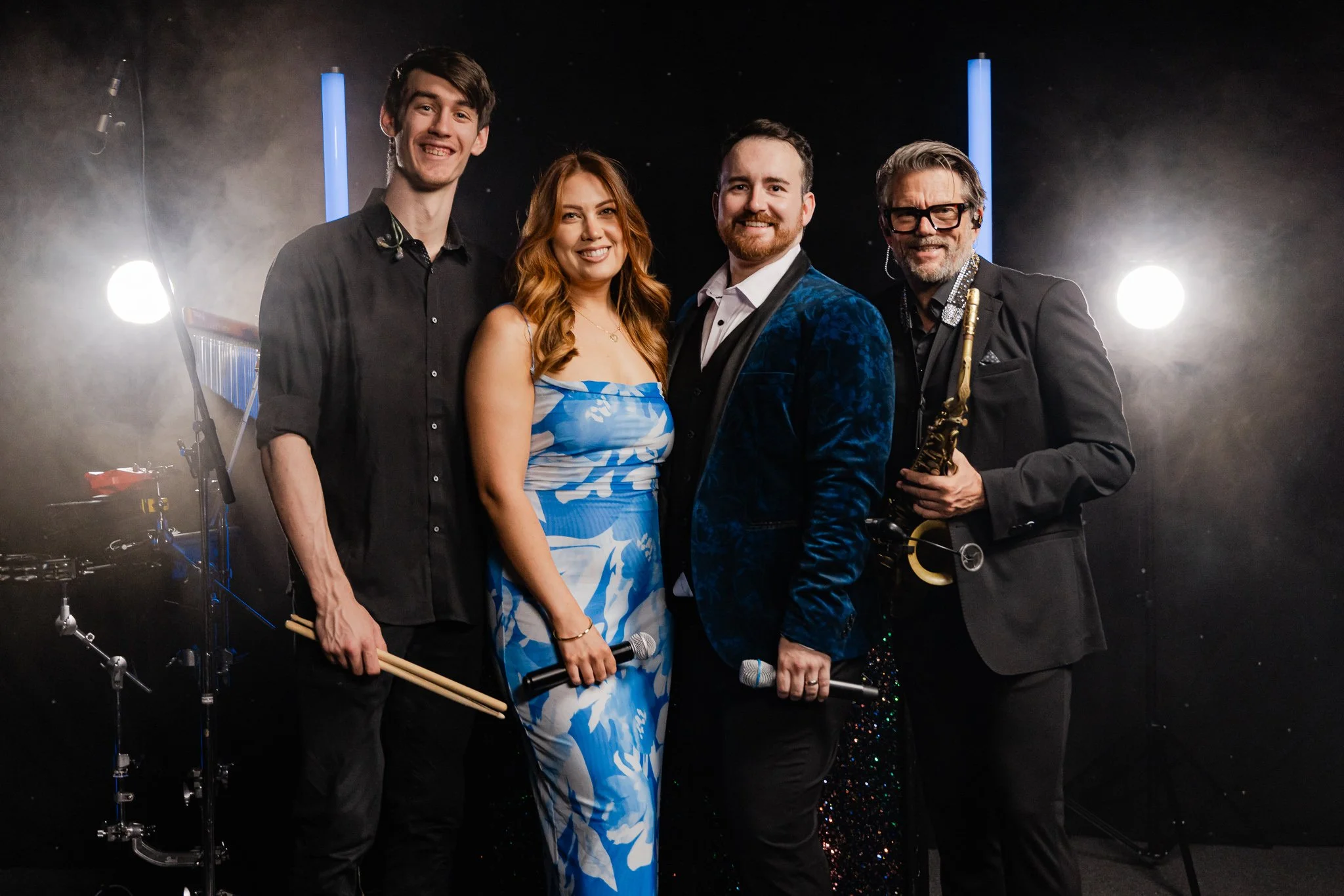 A group of four musicians on a stage with musical instruments, dressed in formal and stylish clothing, smiling at the camera. The background features stage lights and musical equipment.