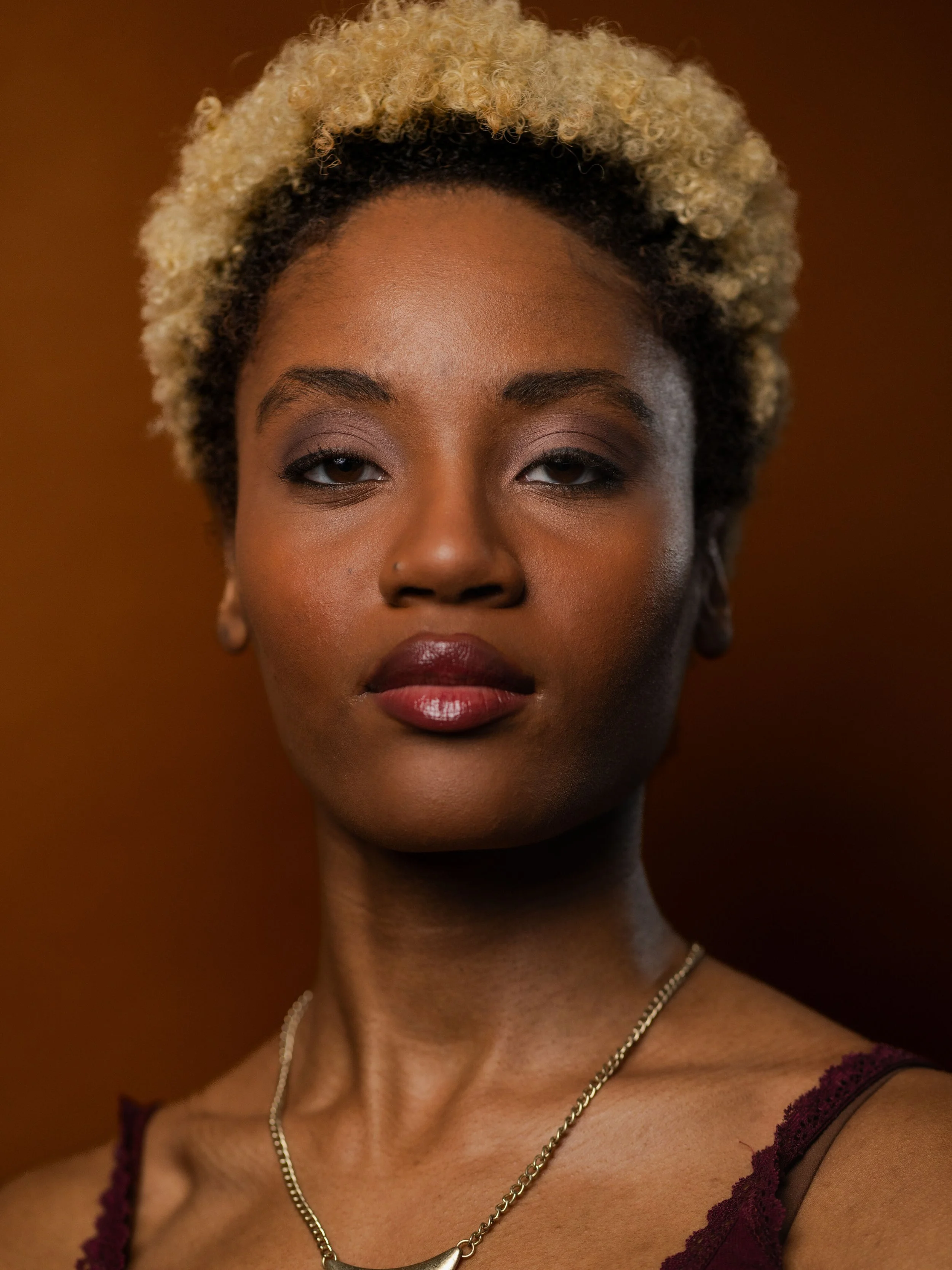Close-up of a woman with curly blonde hair and medium brown skin, wearing a burgundy lace-trimmed top, a silver necklace, and earrings, against a brown background.