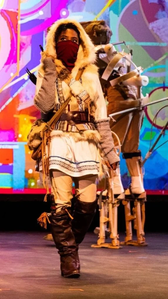 Person dressed in traditional Inuit winter clothing, standing on stage with a colorful background, and a puppet in the background.