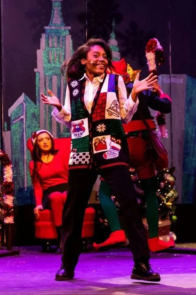 A woman with long dark hair dancing on stage, wearing a festive vest with holiday symbols, black pants, and boots, with a woman sitting in a red chair and holiday decorations in the background.