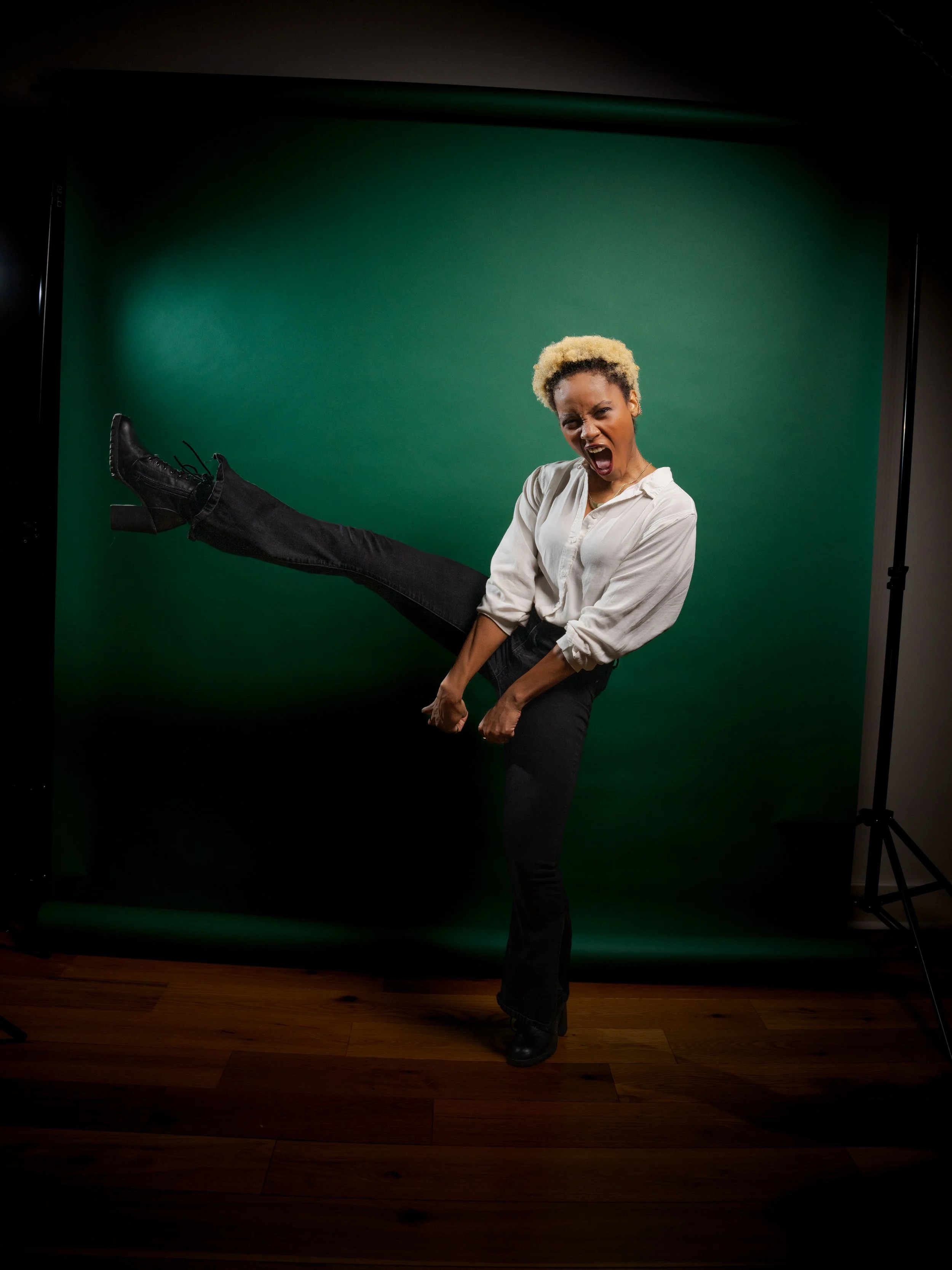 A woman with short blonde hair, wearing a white shirt and black pants, is standing in front of a green screen, yelling or expressing excitement with her mouth open and one leg extended.