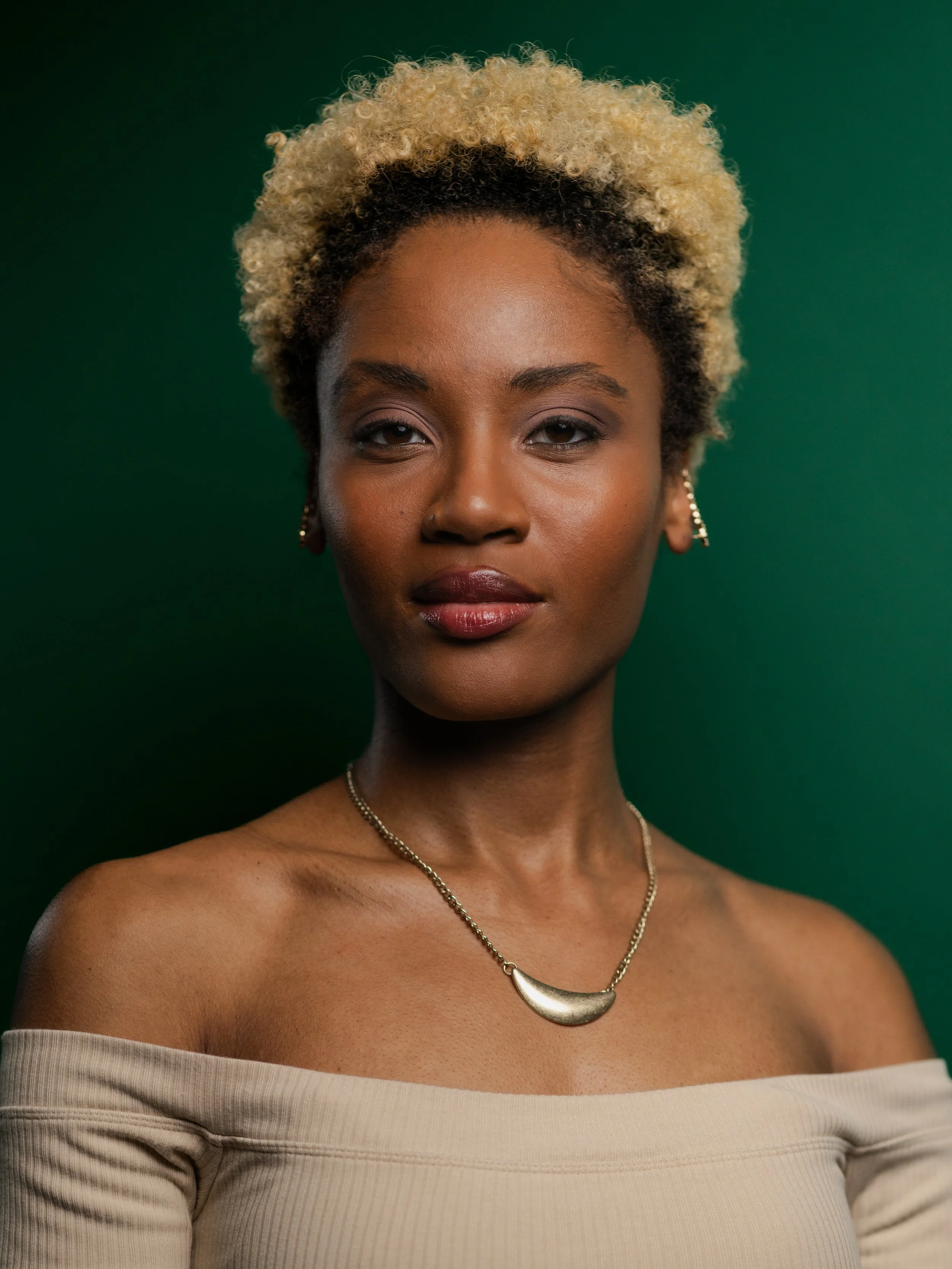 Close-up of a woman with short curly blonde hair, wearing a beige off-the-shoulder top, a silver necklace, and earrings, with a green background.