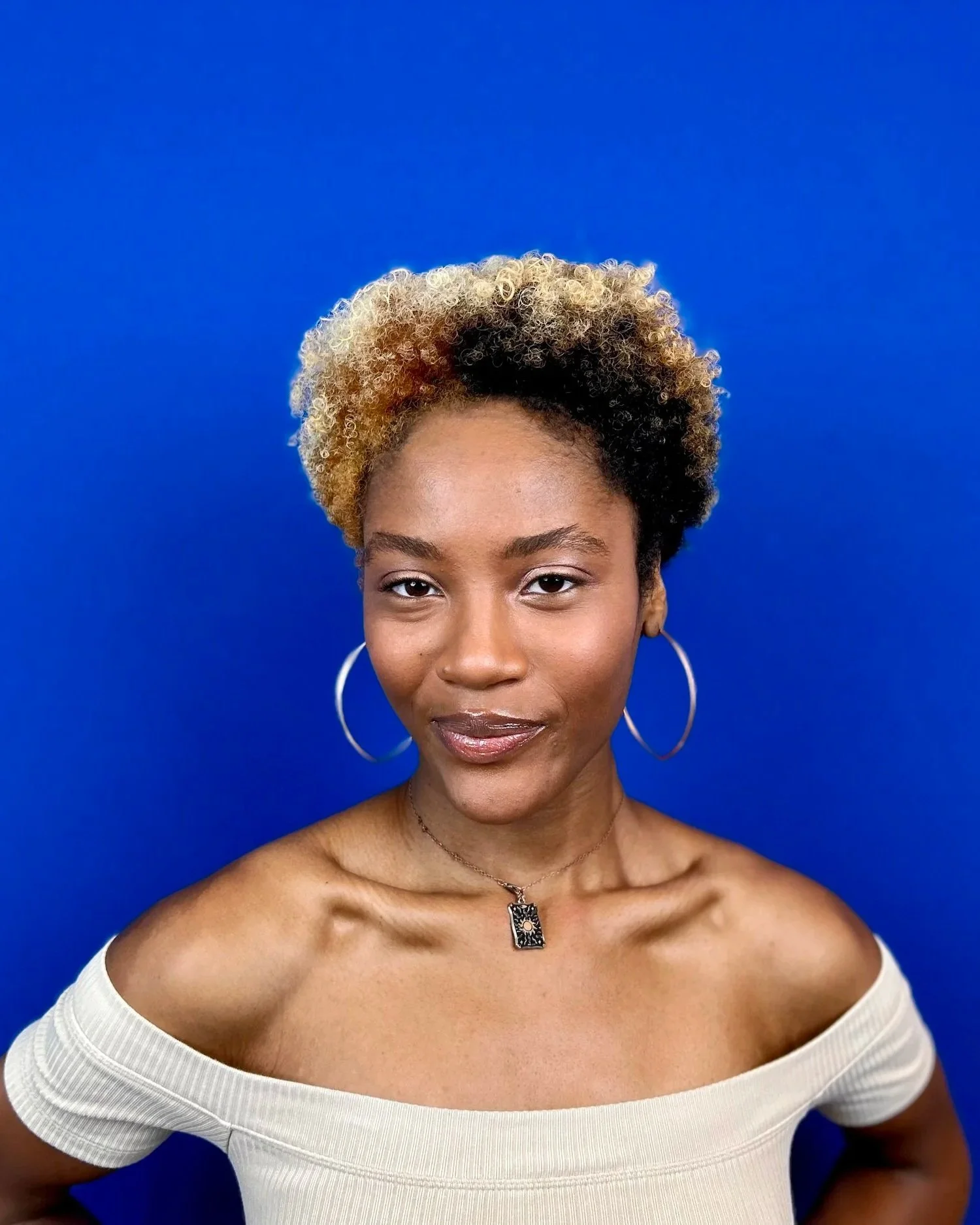 A woman with short, curly hair that is blonde on top and black on the sides, wearing hoop earrings, a necklace with a pendant, and a white off-the-shoulder top, standing against a vibrant blue background.
