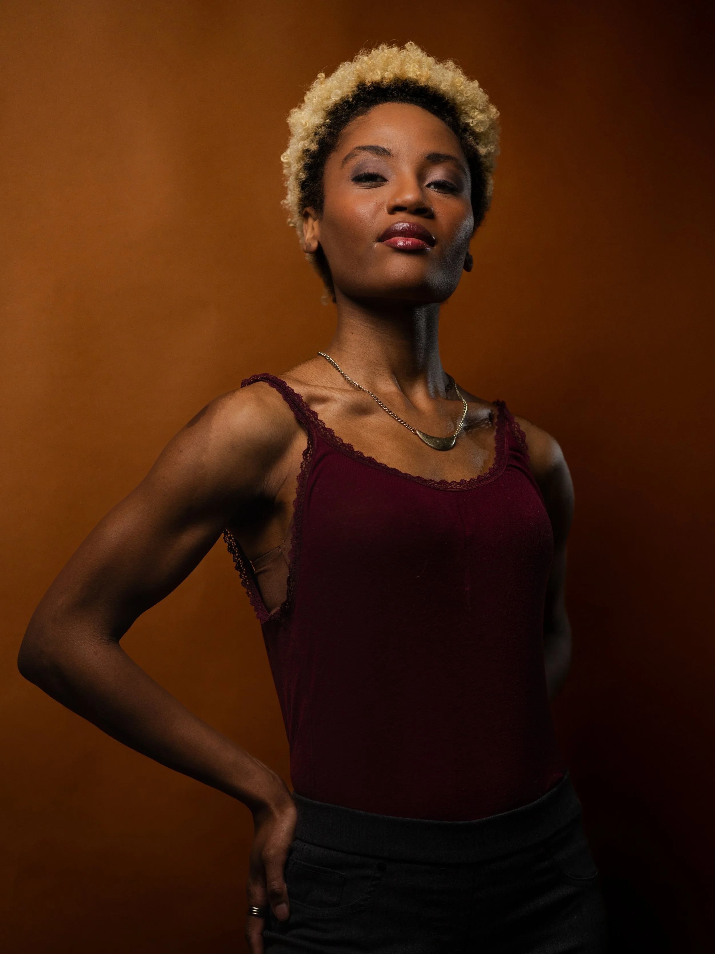 A confident woman with short curly blonde hair, wearing a maroon tank top with lace, black jeans, and a necklace, posing against a brown background.