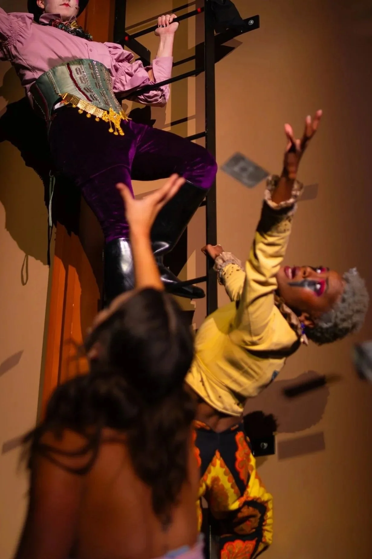 A lively scene of a woman with curly gray hair in a yellow jacket, smiling and gesturing passionately while engaged with a group of people, including children, in an indoor setting.