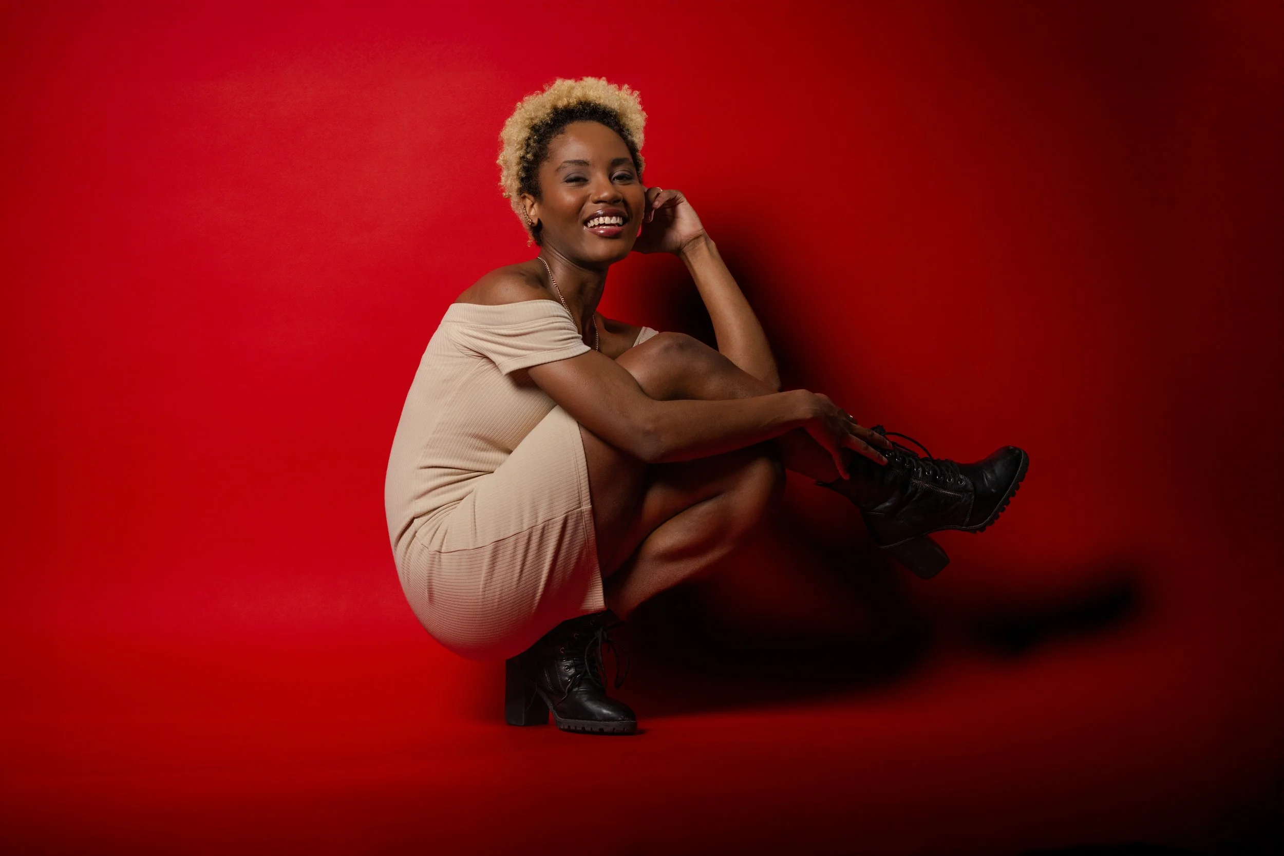 A woman with short, curly blonde hair, wearing a beige off-the-shoulder dress and black combat boots, sitting against a red background, smiling and holding her hair.