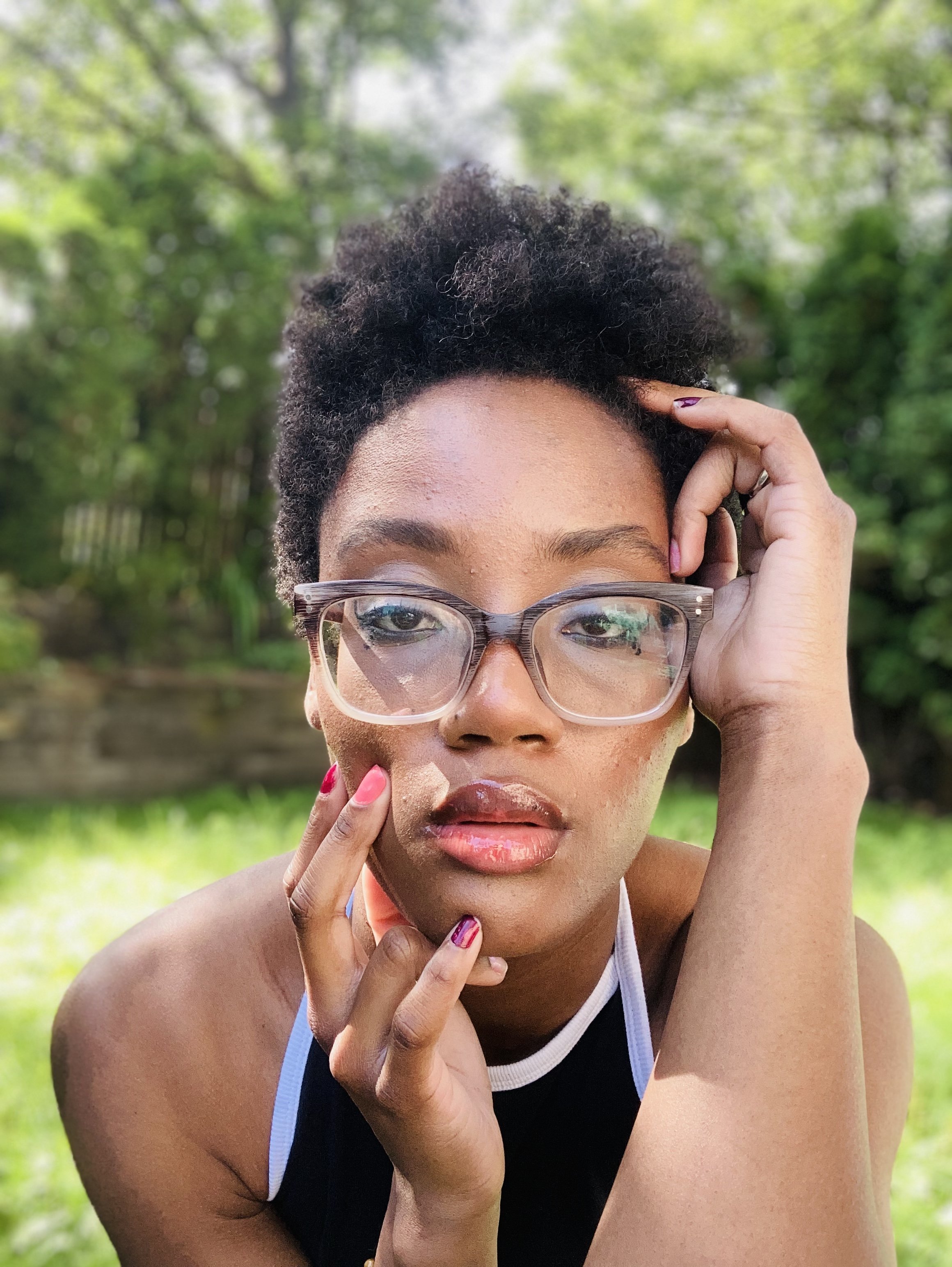 Close-up of a woman with curly black hair wearing glasses and a black and white athletic tank top, holding her chin and face outdoors with a blurred green foliage background.