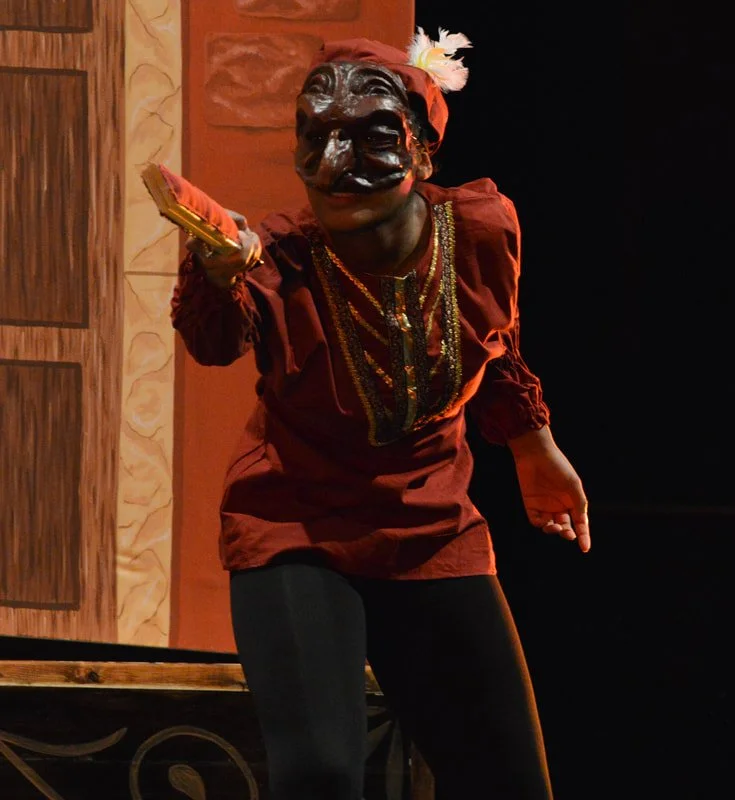 Performer wearing a dark mask, red costume with gold trim, and feathered headpiece, holding a gold object and gesturing with one hand on stage