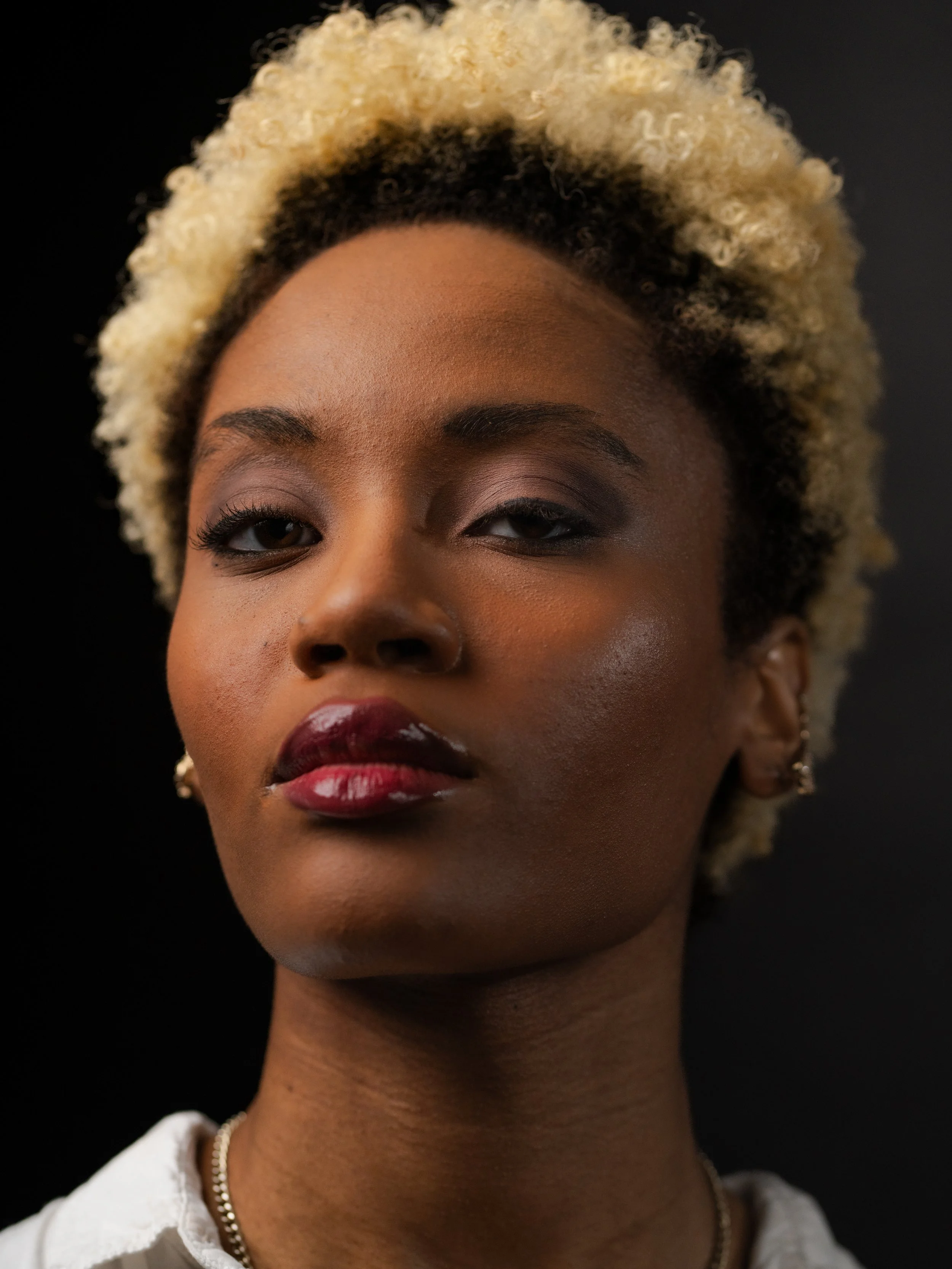 Close-up of a woman with blonde curly hair, dark eyebrows, wearing dark lipstick, earrings, a necklace, and a white shirt, against a black background.