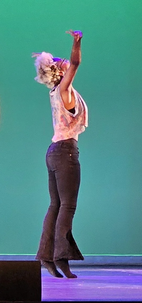A woman with blonde curly hair wearing glasses, a tie-dye tank top, and black bell-bottom pants, standing on stage with a green backdrop, raising one arm in the air.