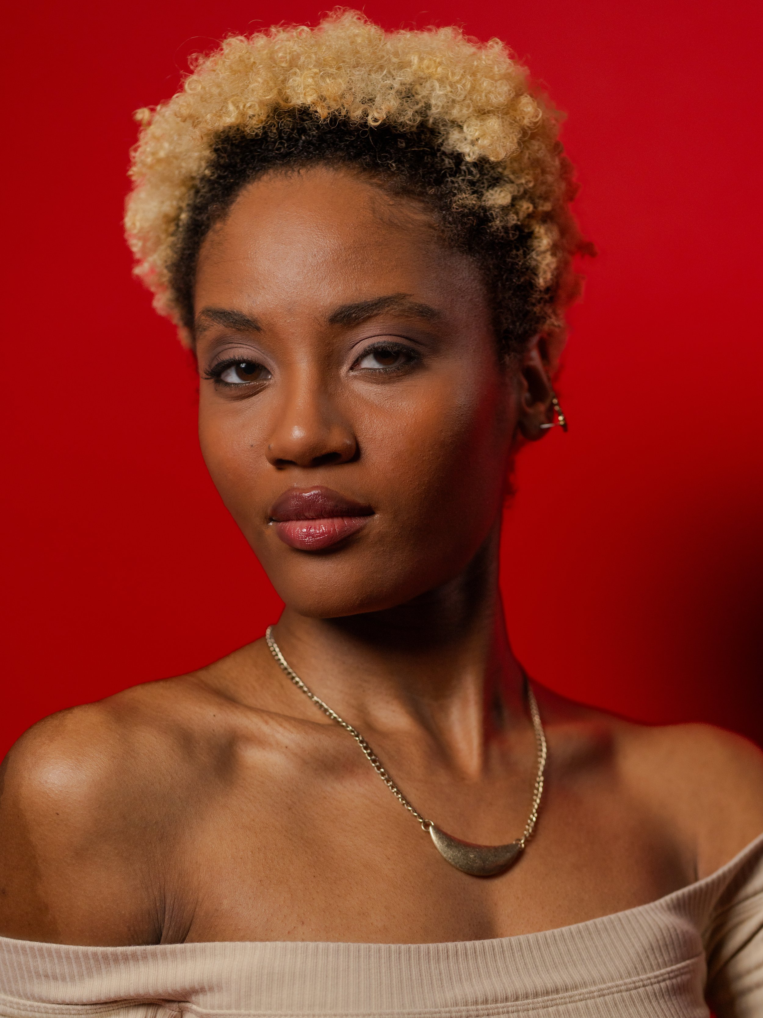 A woman with short, curly, blonde and black hair, wearing makeup, a pearl necklace, earrings, and a beige off-shoulder top, against a red background.