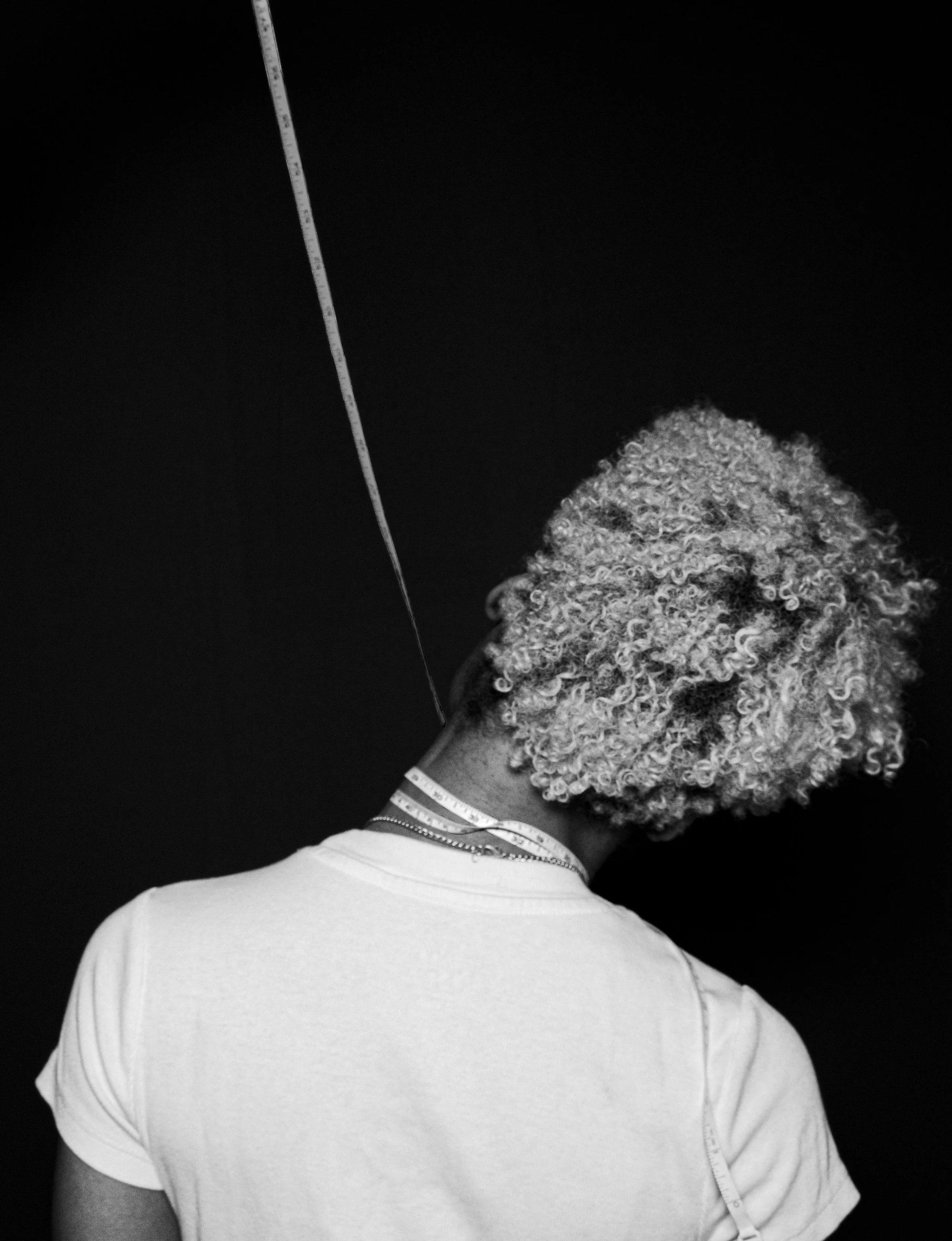 Back of a person with curly hair wearing a white t-shirt, measuring tape around the neck and shoulder, with a dark background.