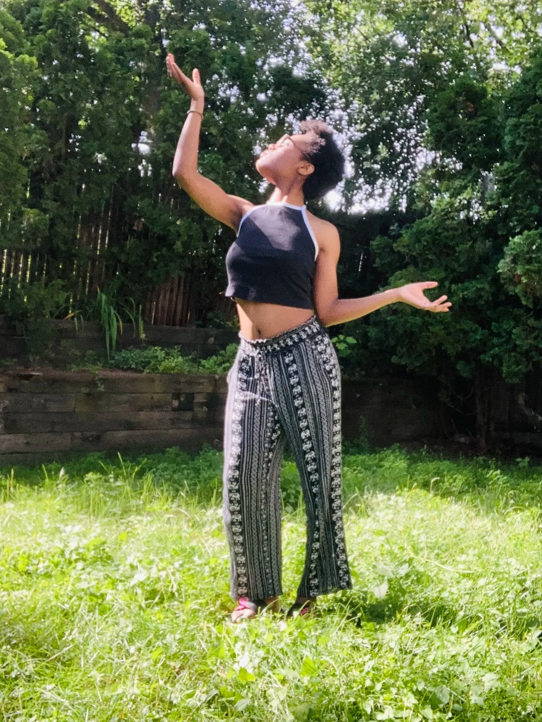 A woman with short curly hair and glasses standing outdoors on grass, wearing a sleeveless top and patterned wide-leg pants, with her hands raised in a joyful pose amidst greenery.