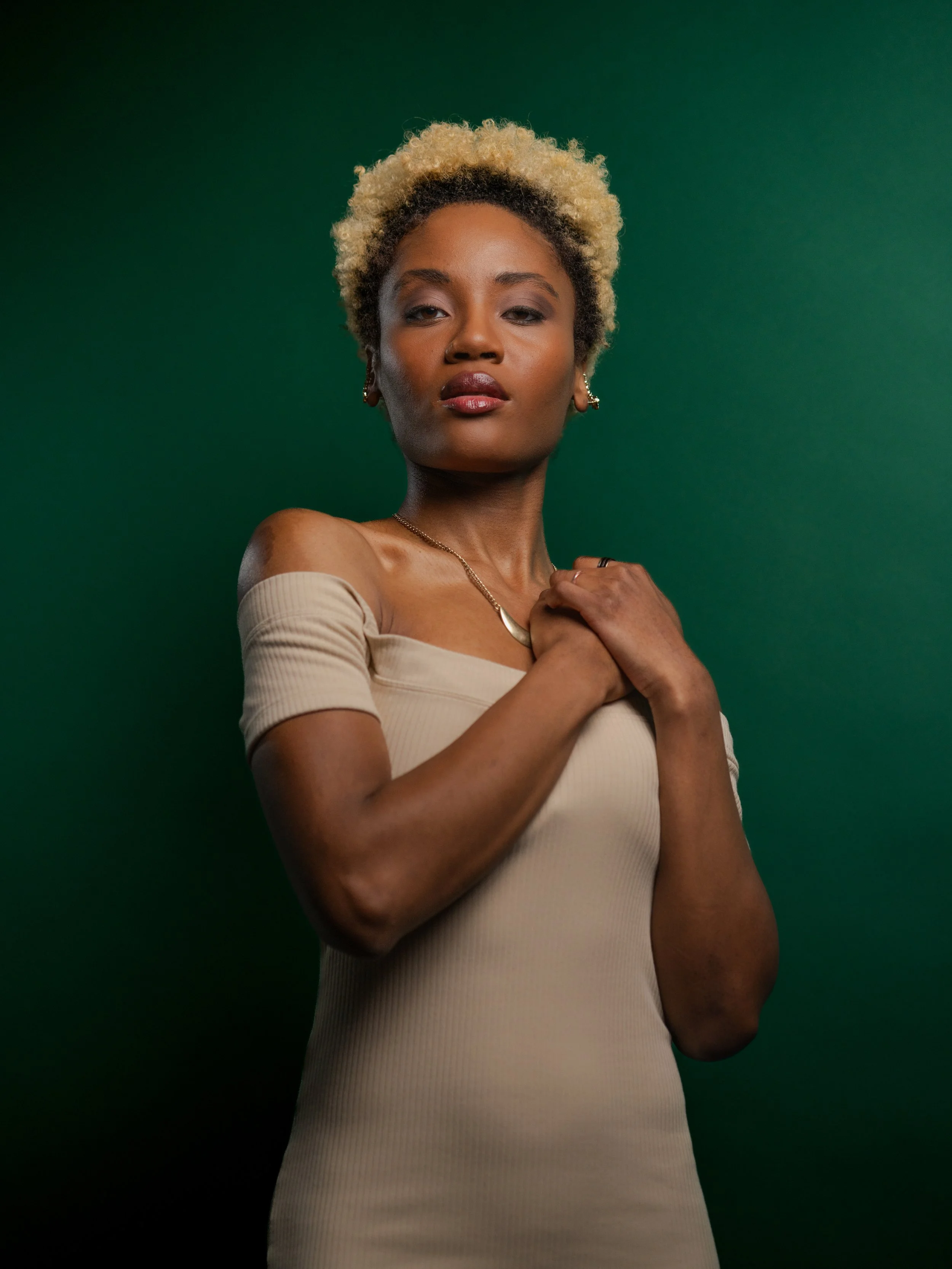 Portrait of a young woman with curly blonde hair, wearing a beige off-shoulder top, earrings, and a necklace, against a dark green background.
