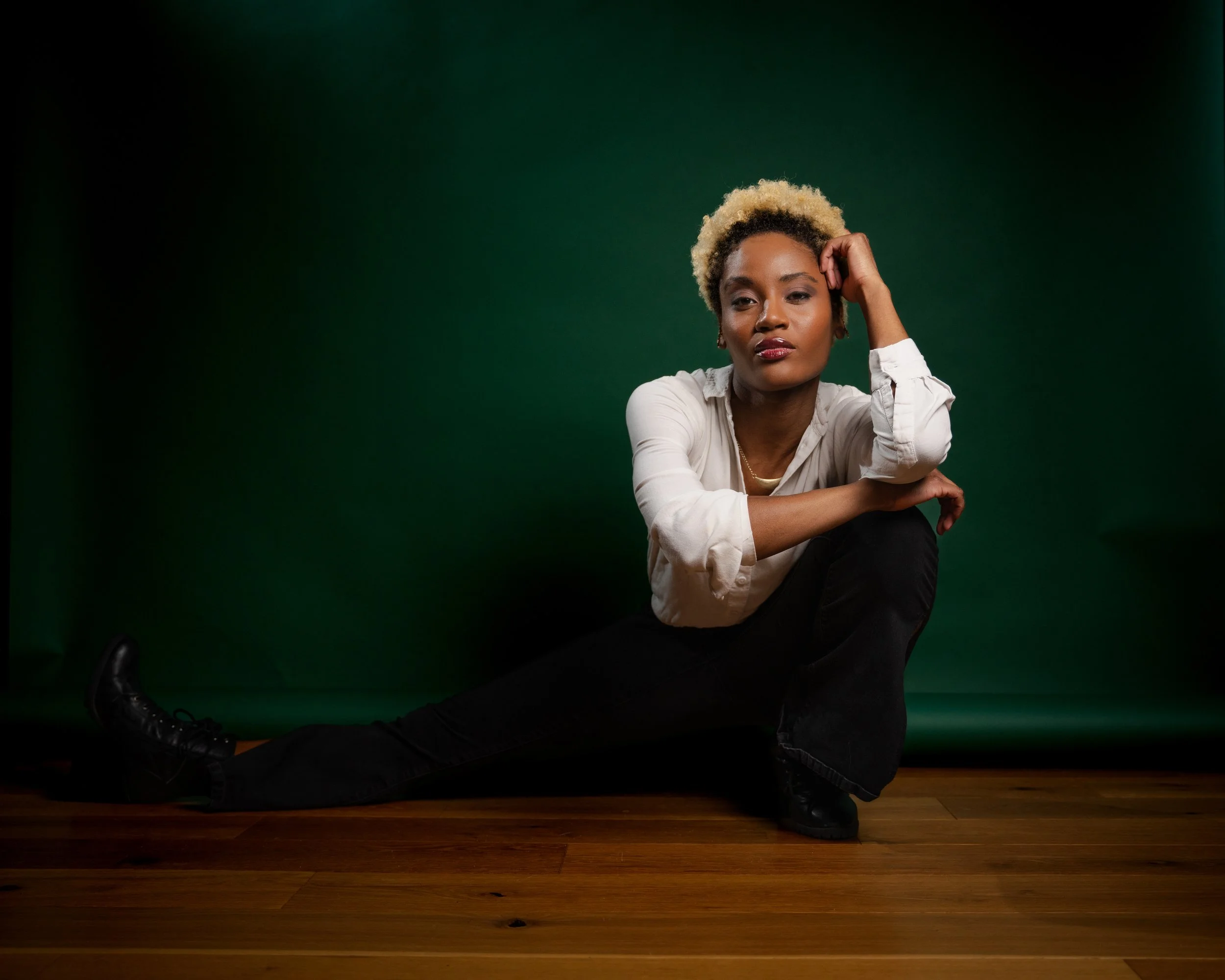 A woman with short blonde hair and dark skin, wearing a white shirt and black pants, is sitting on a wooden floor in front of a green background. She poses with her left hand resting on her head and her right arm across her lap, looking at the camera with a confident expression.