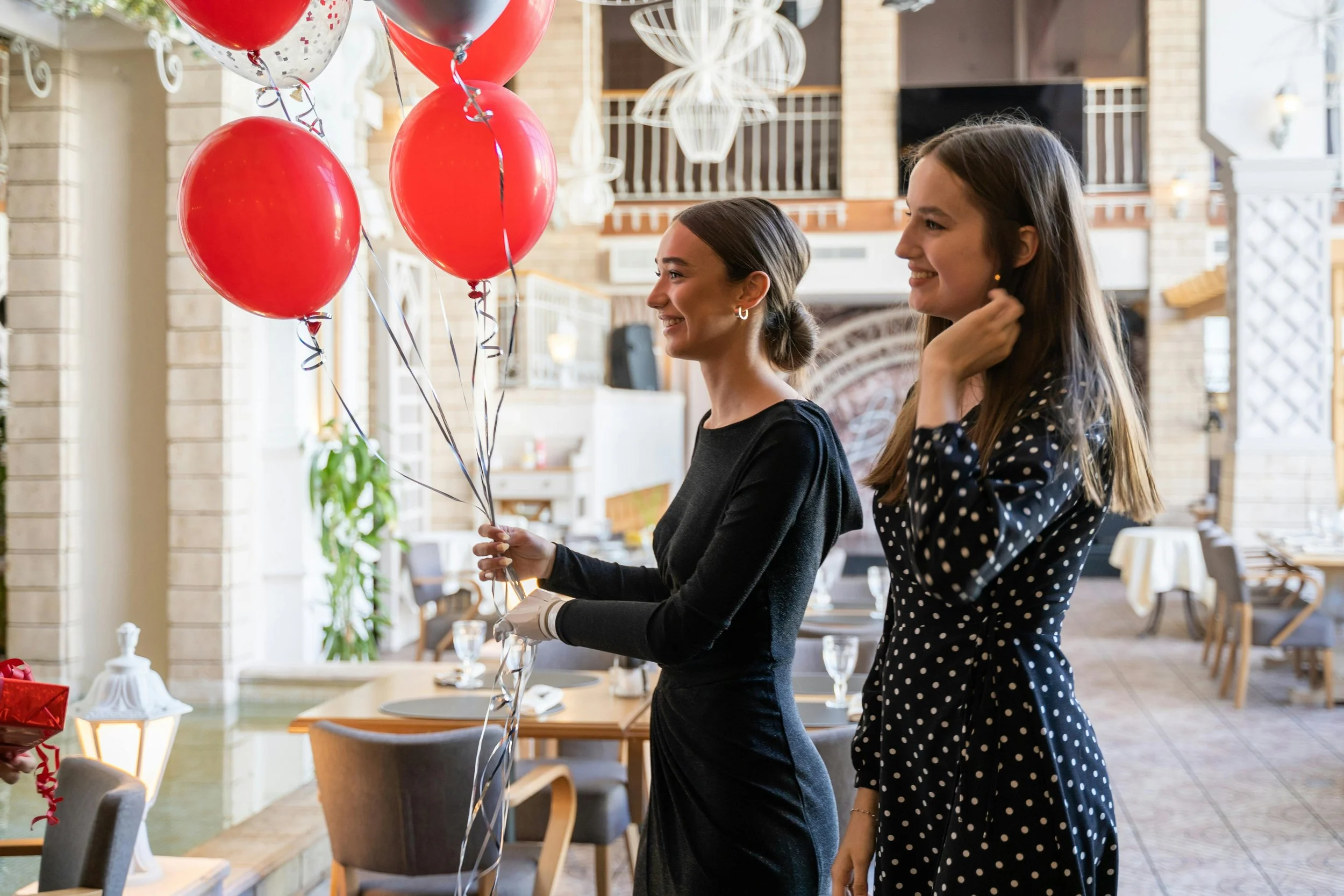 Two young women smiling during a celebration in a decorated restaurant, one woman holding red balloons, the other wearing a polka dot dress.