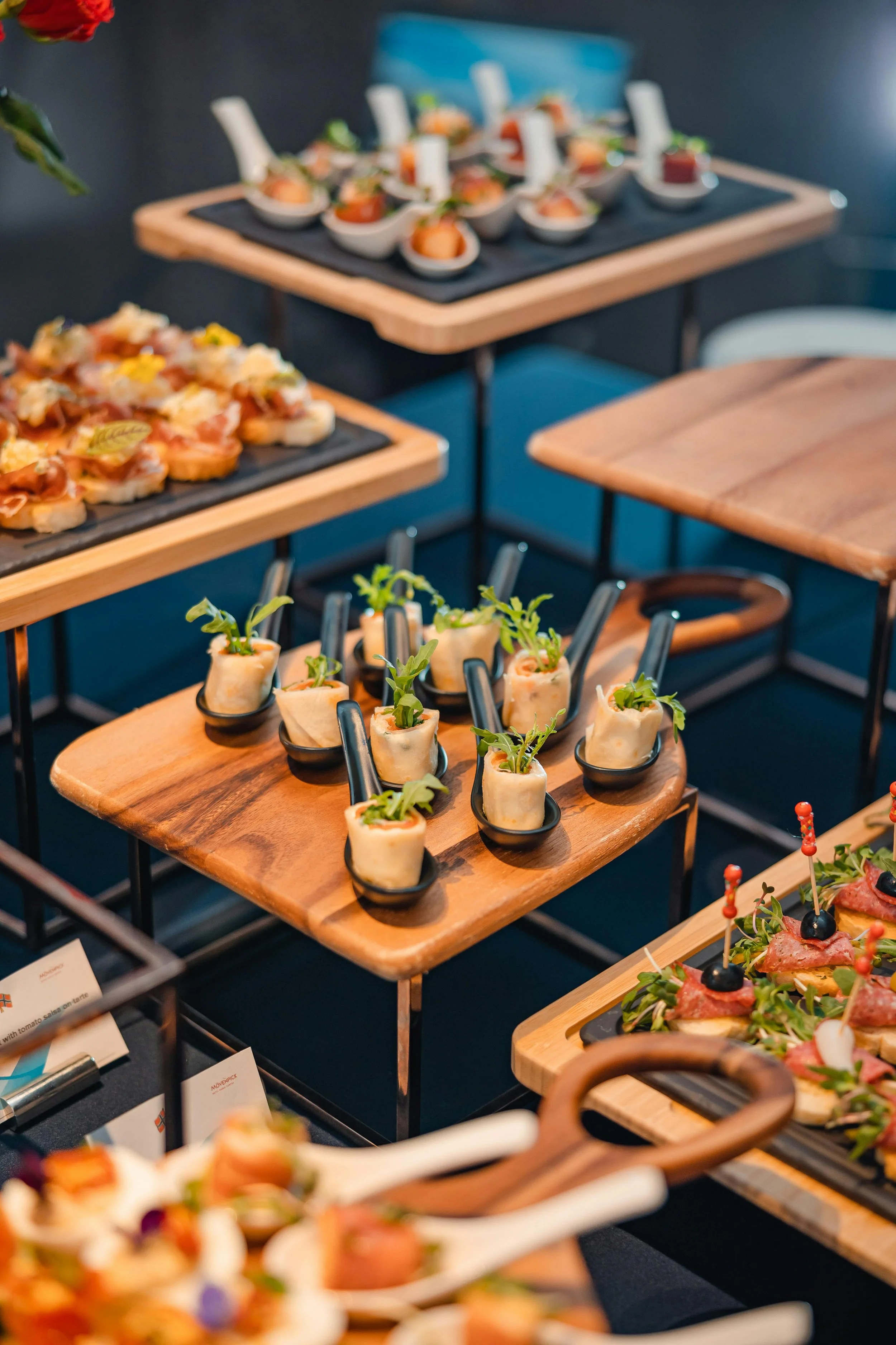 Assorted appetizers on wooden and black platters, including mini spring rolls with green garnish, sliders with cured meat and cheese, and various finger foods, set on a table.