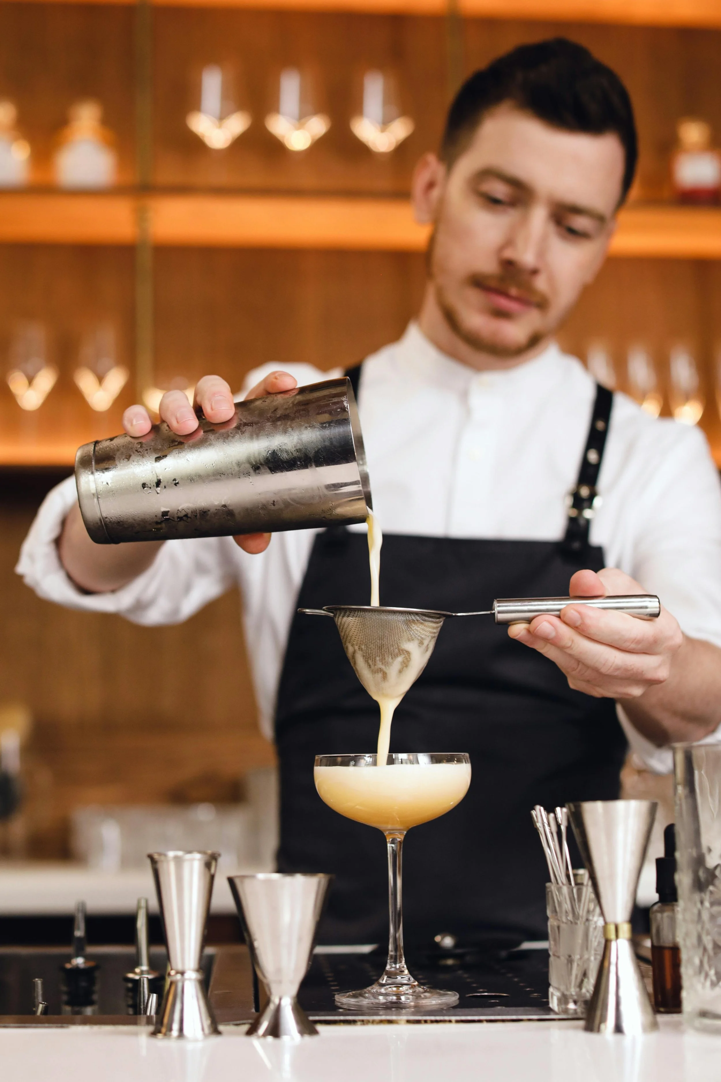 A bartender pours a foamy yellow cocktail through a strainer into a coupe glass.