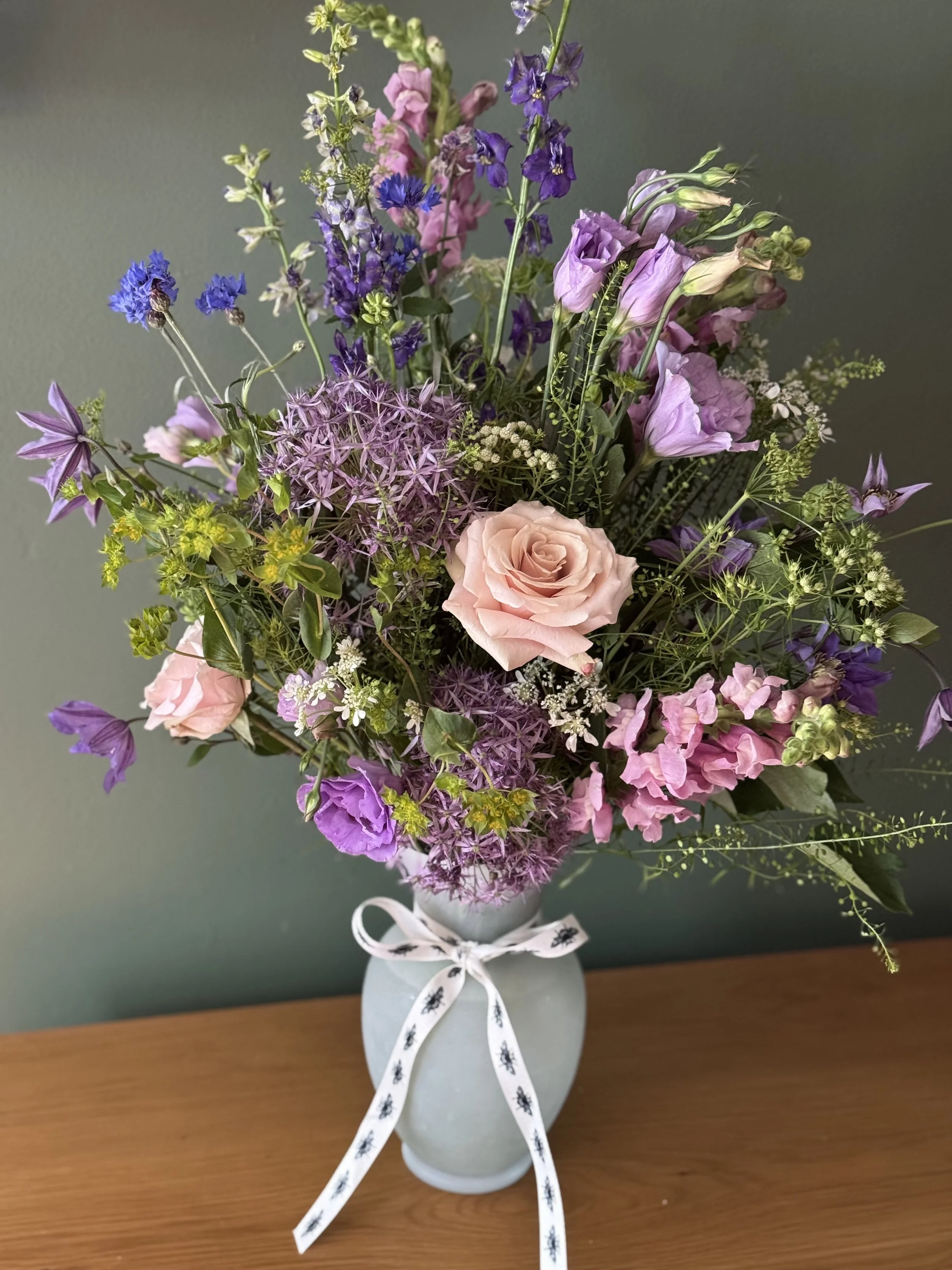 A bouquet of mixed pink, purple, and lavender flowers in a light gray vase with a white ribbon tied around it, placed on a wooden surface against a gray background.