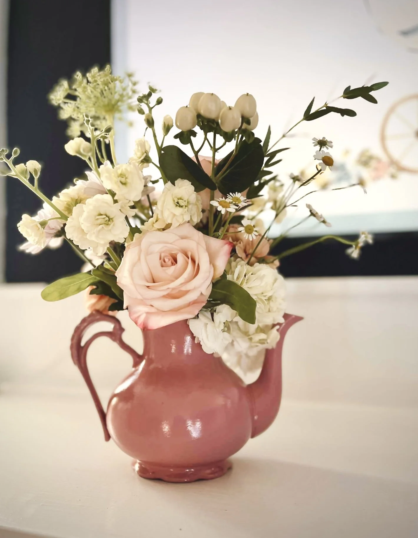 A pink ceramic pitcher containing a bouquet of white, pink, and green flowers and greenery, placed on a white surface with a blurred background.