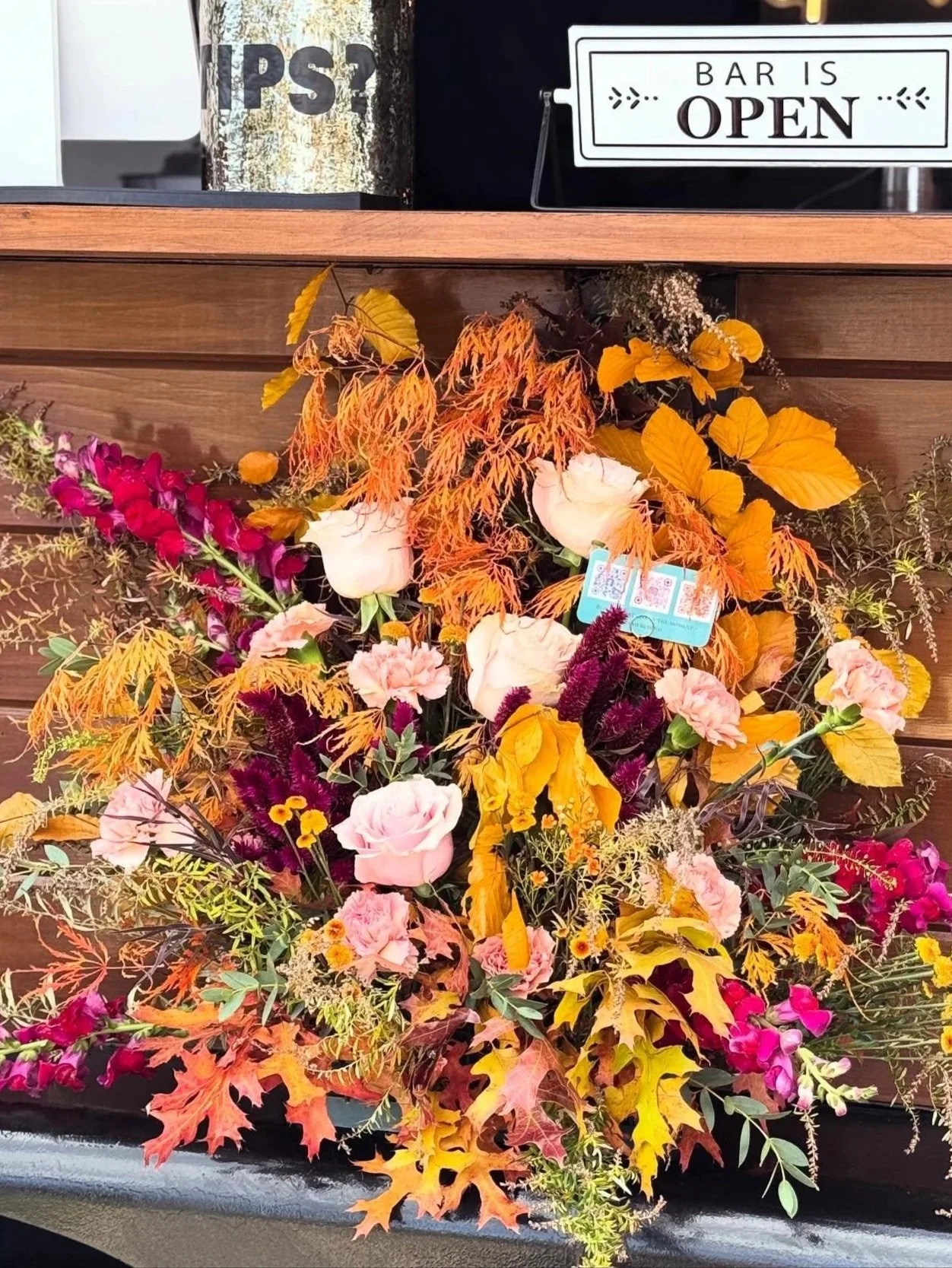 A floral display with pink roses, purple and yellow flowers, and orange and yellow autumn leaves, situated on a black surface against a wooden background. Above, there are signs indicating a bar is open and a metallic container with partially visible text.