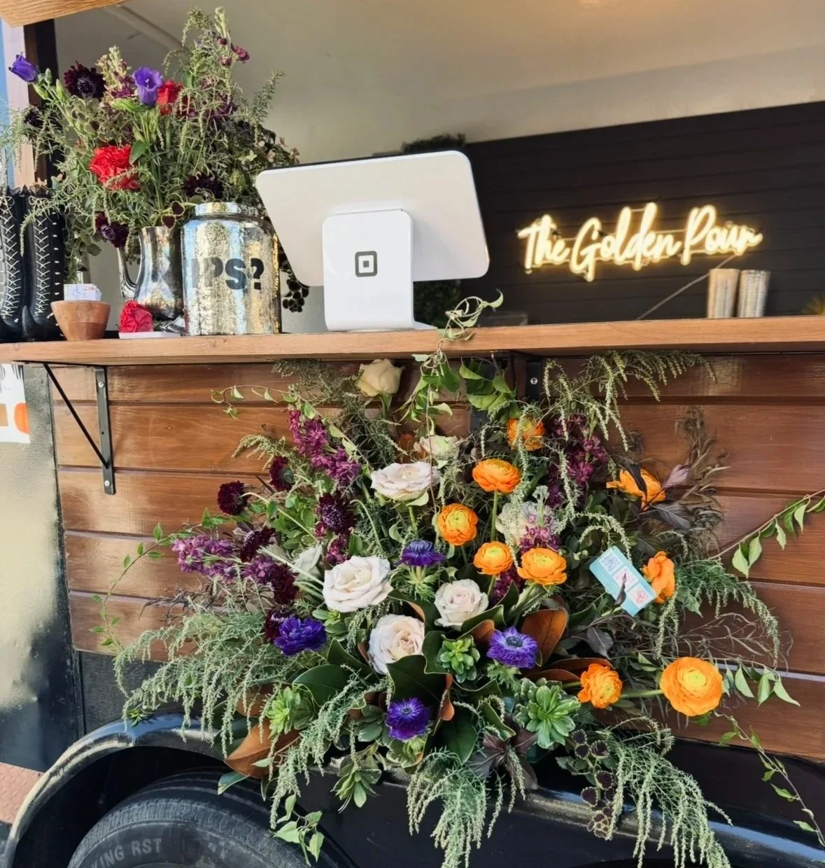 A flower cart with a wooden side, decorated with a large arrangement of colorful flowers, parked in front of a building with a neon sign that reads 'The Golden Pear.' The cart has a cash register on top and is set against a black wall behind the sign